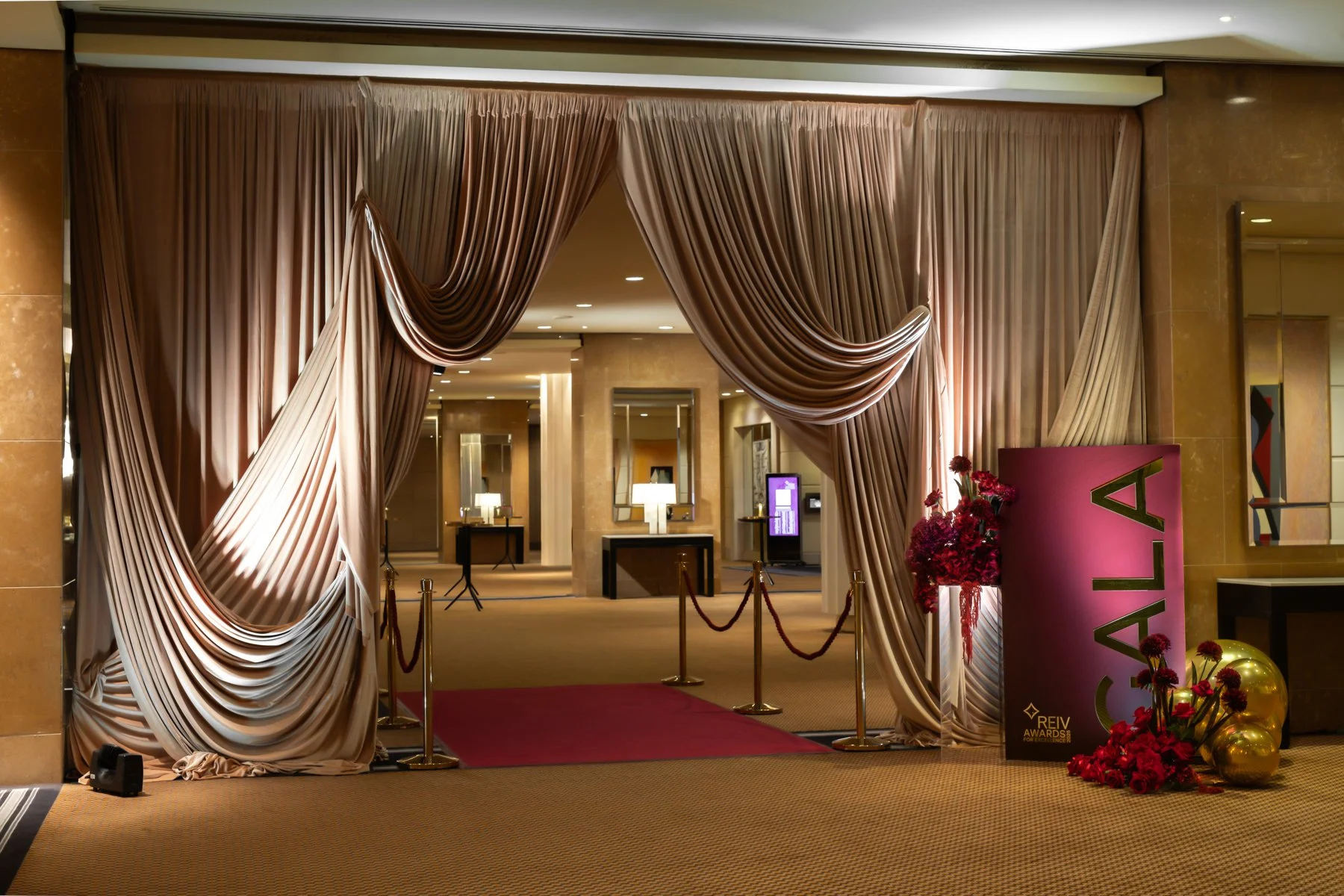 Elegant hotel lobby entrance decorated with large satin curtains, a red carpet, flowers, and a sign that reads "Catal" for the REIV Awards, with warm lighting and modern decor.