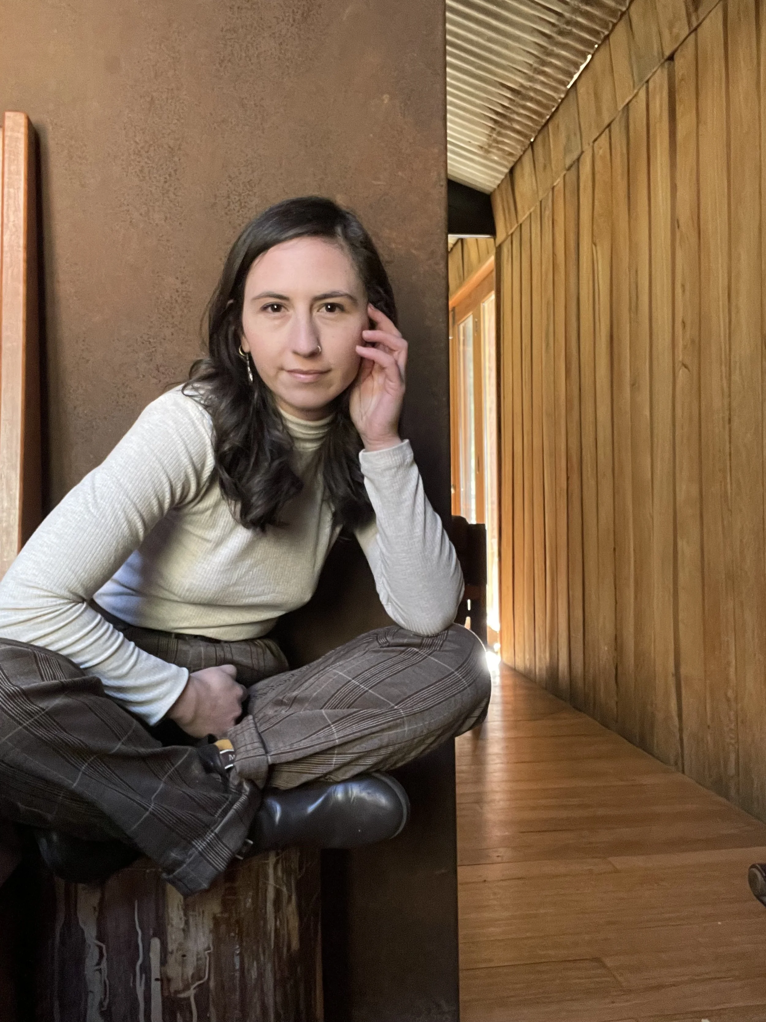 A young woman with dark wavy hair and a nose piercing sitting cross-legged on a wooden surface in a cozy room with wooden walls and ceiling, wearing a beige long-sleeve top and plaid trousers.