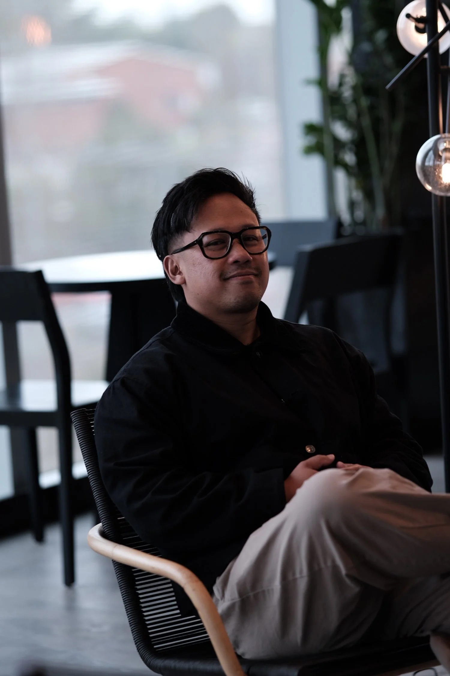 A man with black glasses and dark hair sitting comfortably in a modern cafe or restaurant with large windows and indoor plants.