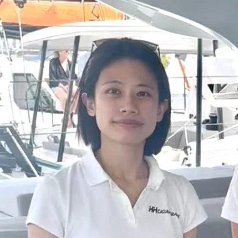A woman with short black hair wearing glasses, a collared white HH Catamarans shirt standing at a boat show booth.