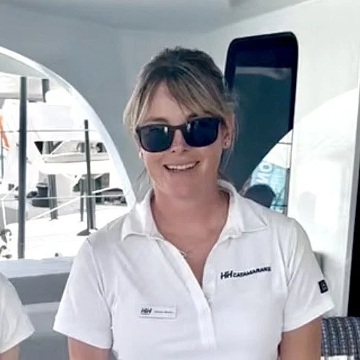 A woman smiling outdoors near a body of water, with sunglasses on her head and a cloudy sky in the background.