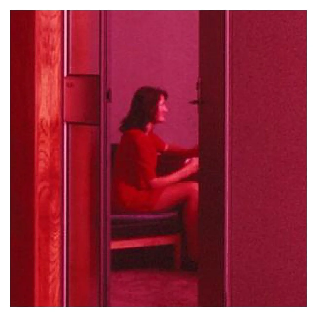 Woman sitting on a bench in a small room with pinkish-red lighting.