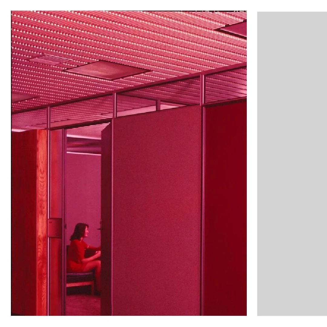 A woman in a red dress sitting in a small booth with red walls in a modern interior space, illuminated by pink lighting and a perforated ceiling.