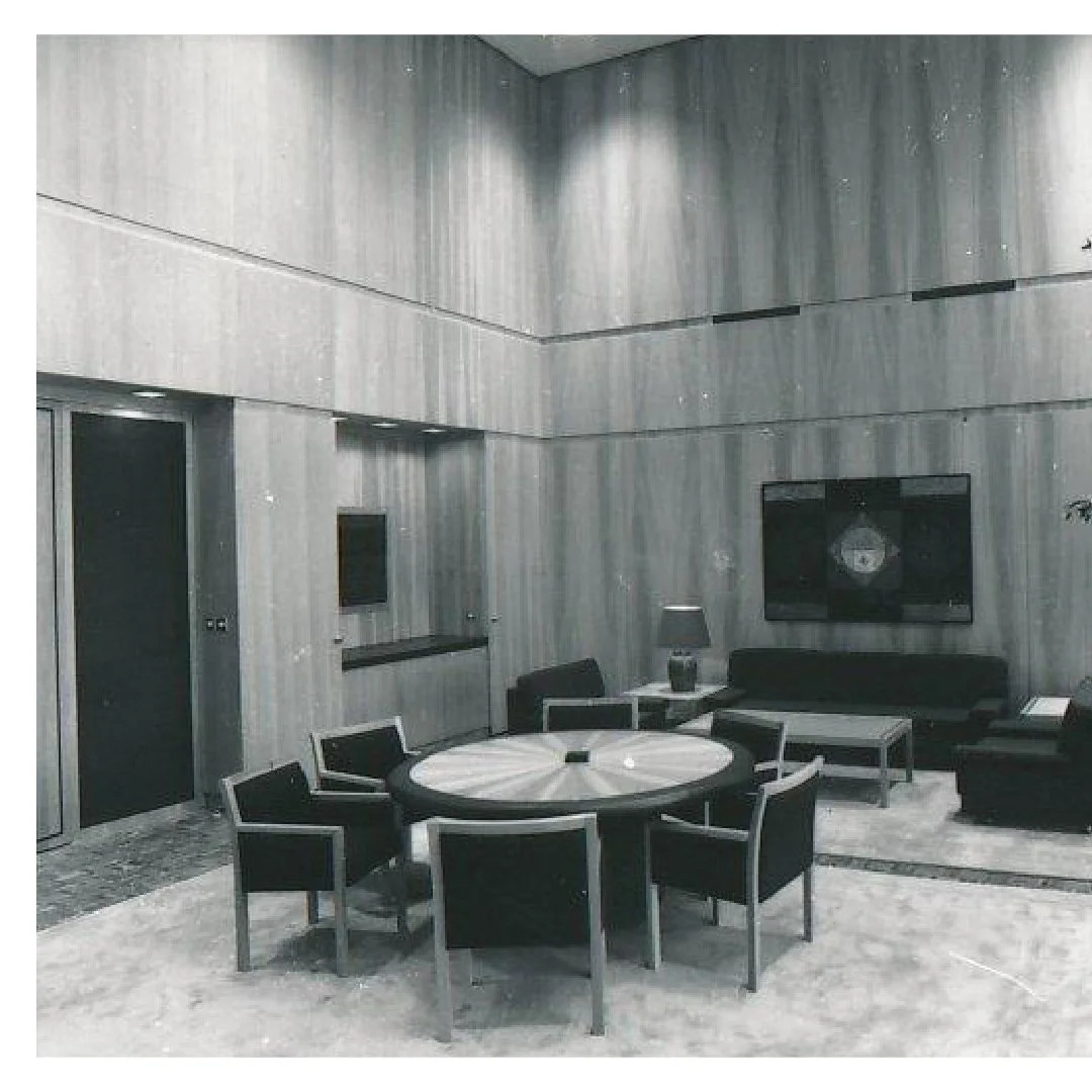 Modern lounge area with a round table surrounded by six black chairs, a black sofa, a side table with a lamp, and a large wall-mounted television in a room with wooden panel walls and marble flooring.