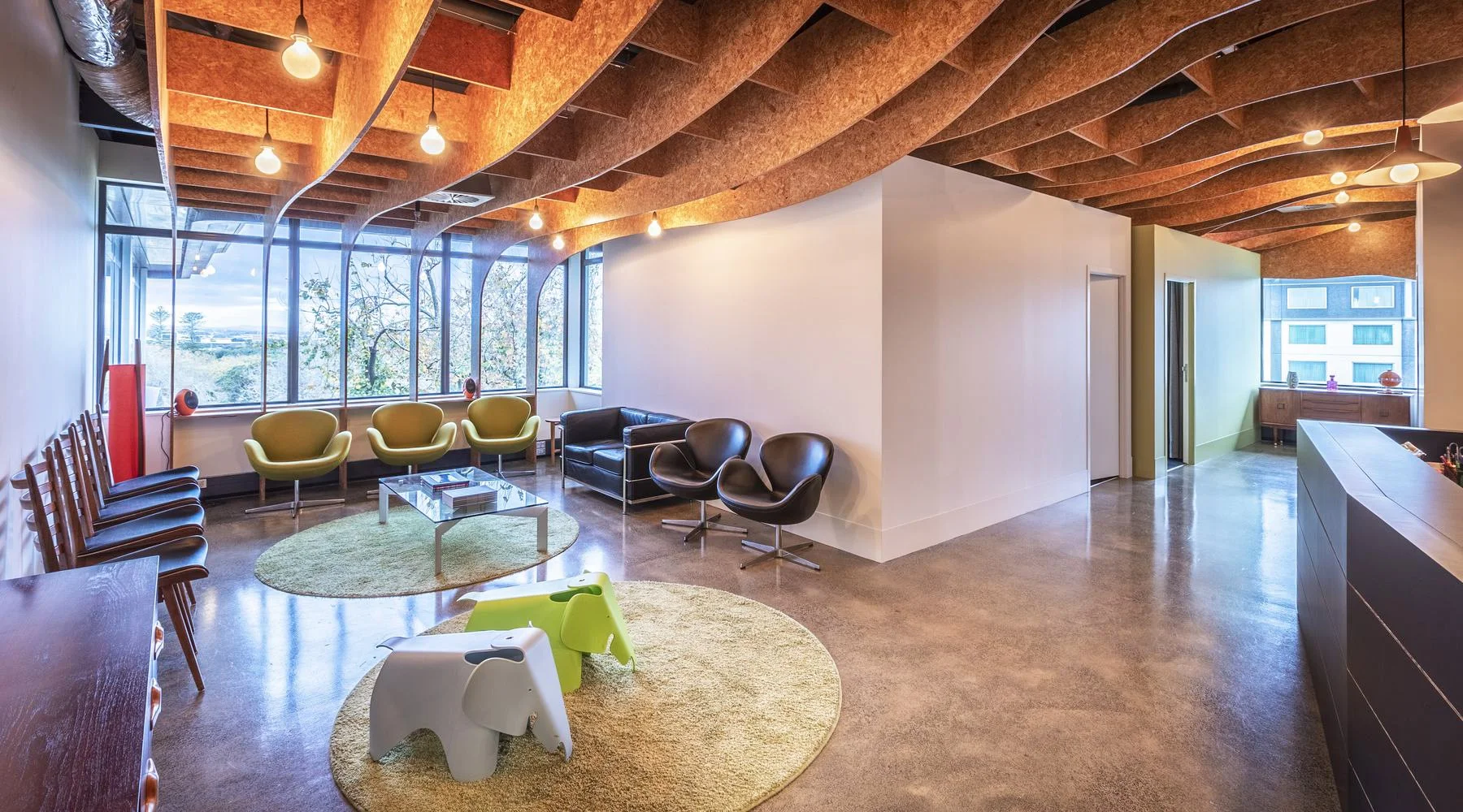 Modern office waiting area with green, black, and wooden chairs, a glass coffee table, and playful elephant-shaped stools, featuring large windows, wooden ceiling design, and minimal decor.