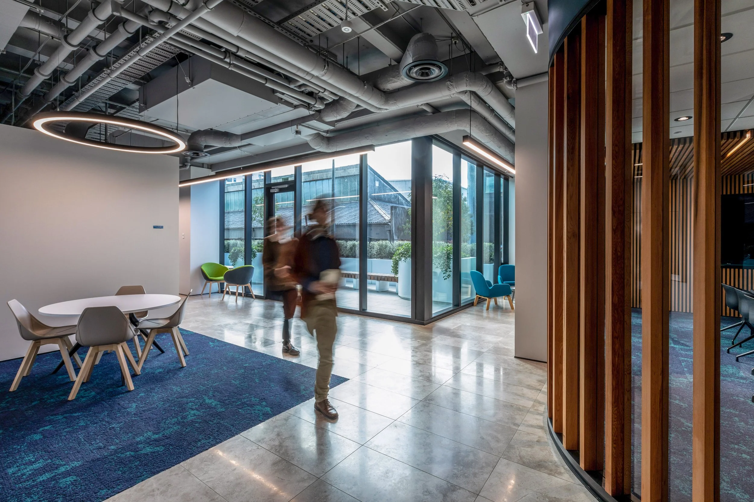 Modern office lobby with glass walls, contemporary furniture, and exposed ceiling pipes. Blurred people walk through the space, which has a mix of seating areas, including chairs and a round table. Natural light floods in from large windows.