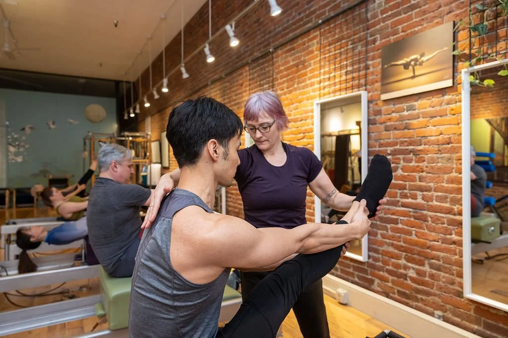 A fitness instructor assisting a man with stretching exercises in a gym with brick walls, mirrors, and other people exercising in the background.