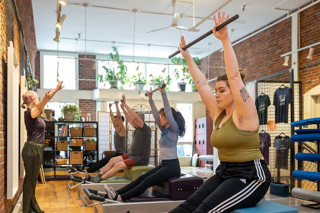 Instructor leading a fitness class with four women exercising with bars in a studio with brick walls and large windows.