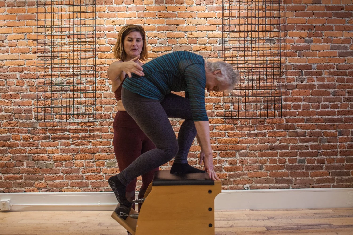 A woman assisting an elderly woman with a fitness exercise on a wooden step platform in a studio with a brick wall background.
