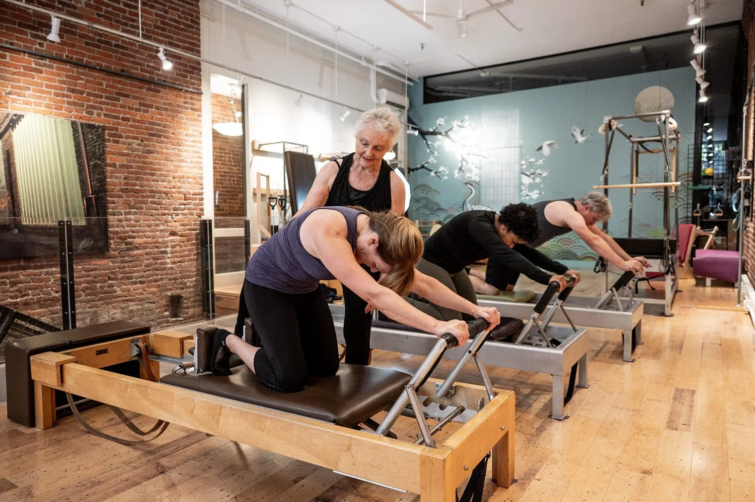 A group of women practicing Pilates on reformer machines in a fitness studio with exposed brick walls and decorative wall art.