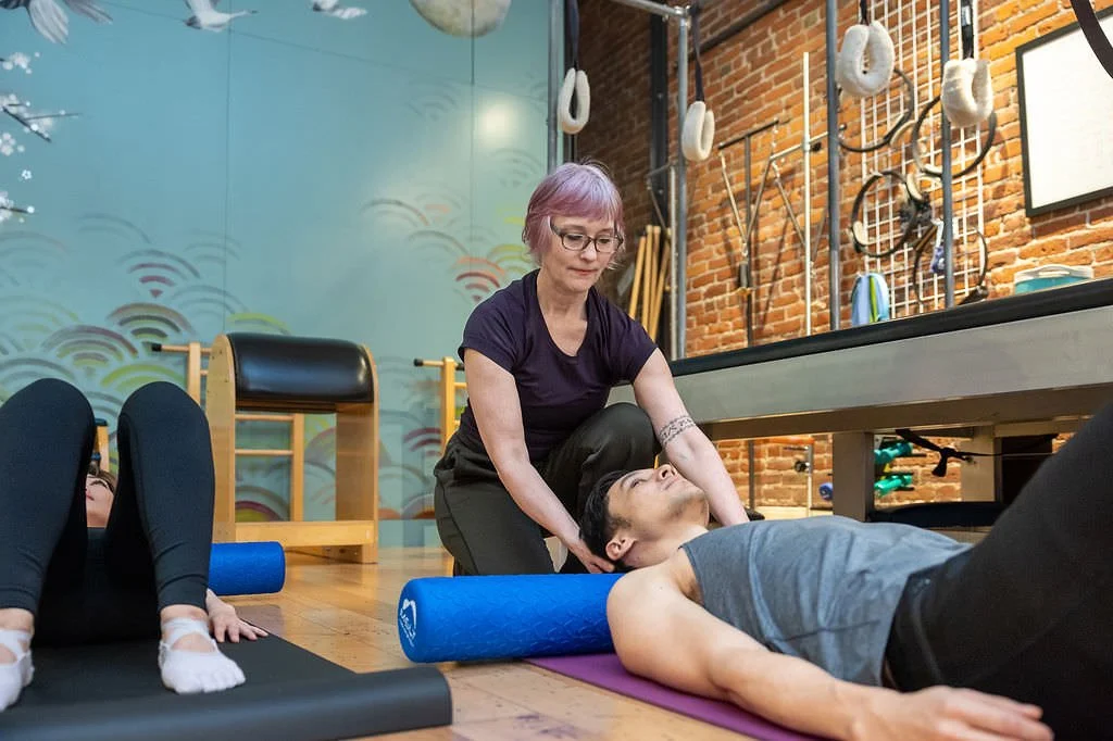 A woman helps a man stretch on a yoga mat in a fitness studio with gym equipment hanging on the brick wall in the background.