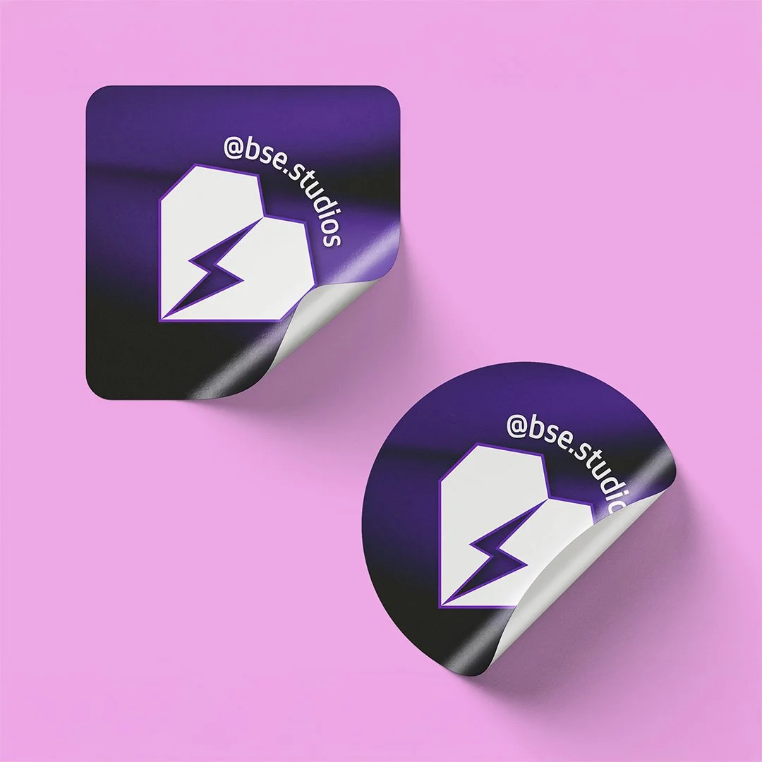 Due adesivi circolari con sfondo in gradiente di viola e nero, raffigurano un cuore con una scarica di energia, con il testo '@bse.studios'.