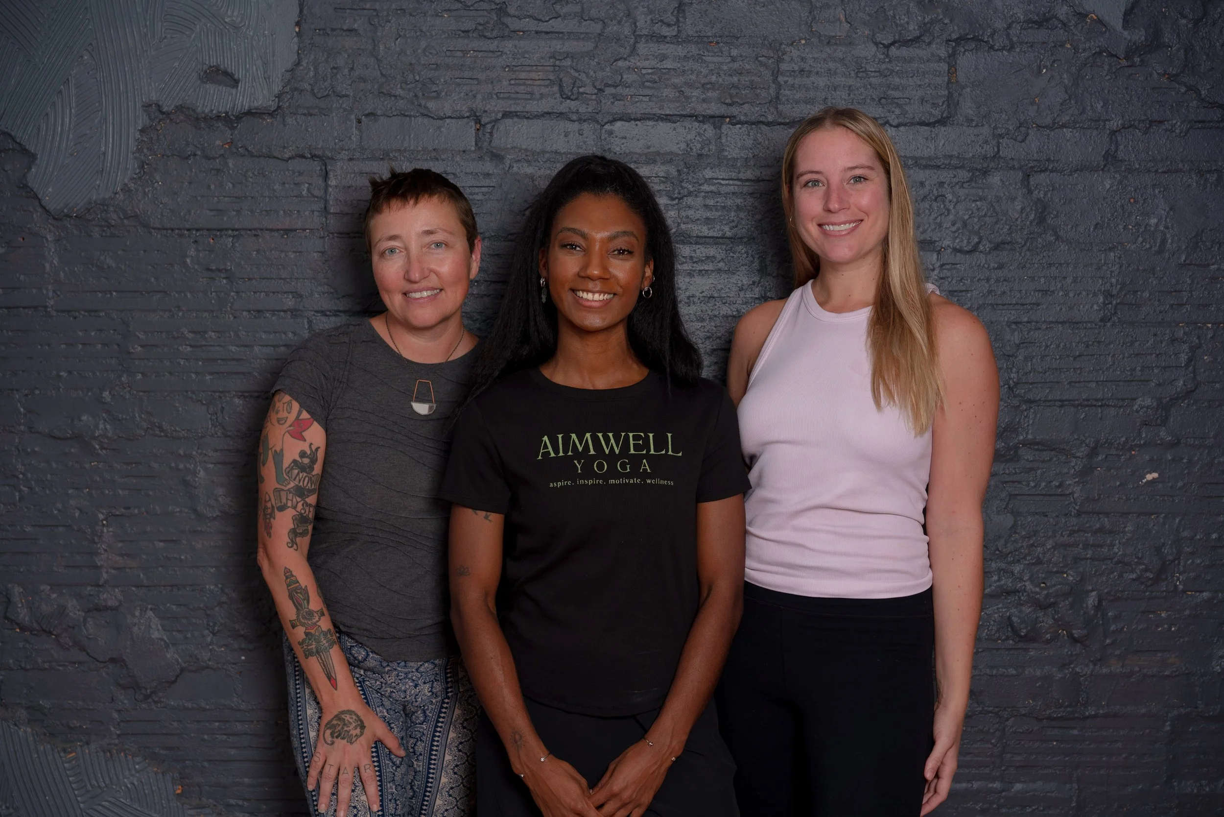 Three women standing side by side against a dark brick wall, smiling at the camera. The woman on the left has short hair, tattoos on her arm, and is wearing a dark gray shirt and patterned pants. The woman in the middle has long black hair, is wearing a black T-shirt with 'AIMWELL YOGA' written on it, and has earrings. The woman on the right has long blonde hair, is wearing a white sleeveless top, and black pants.