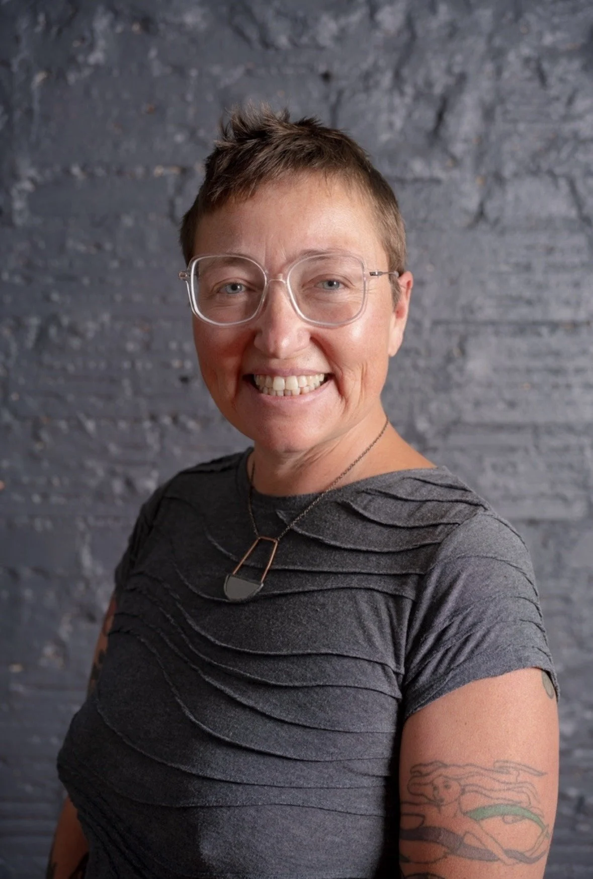 A woman with short hair, glasses, and tattoos on her arm smiling at the camera, wearing a dark gray shirt and a necklace, standing against a gray brick wall.
