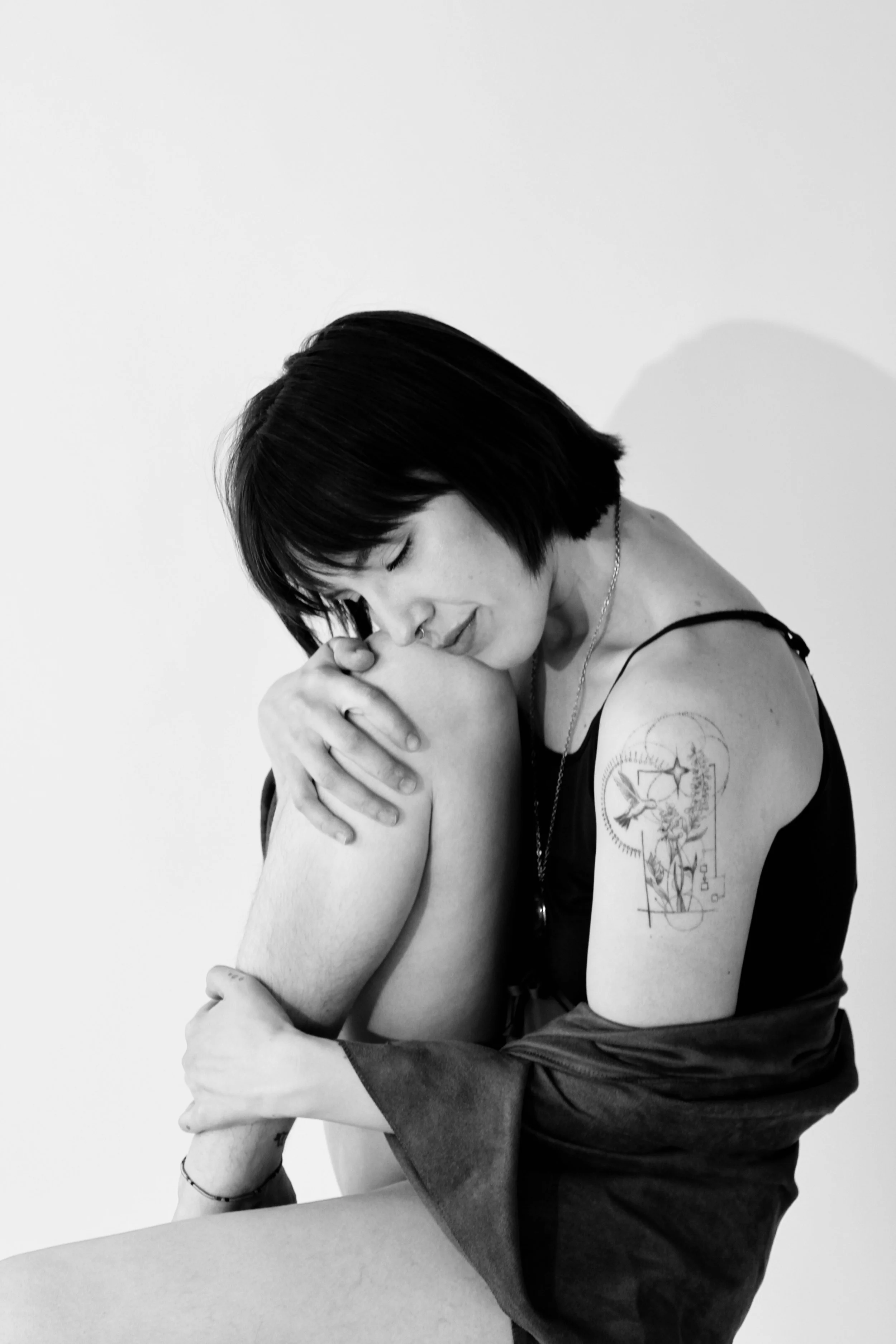 A black and white photo of a woman hugging a person with their face hidden. The woman has short dark hair, visible tattoos on her arm, and is wearing a sleeveless top.