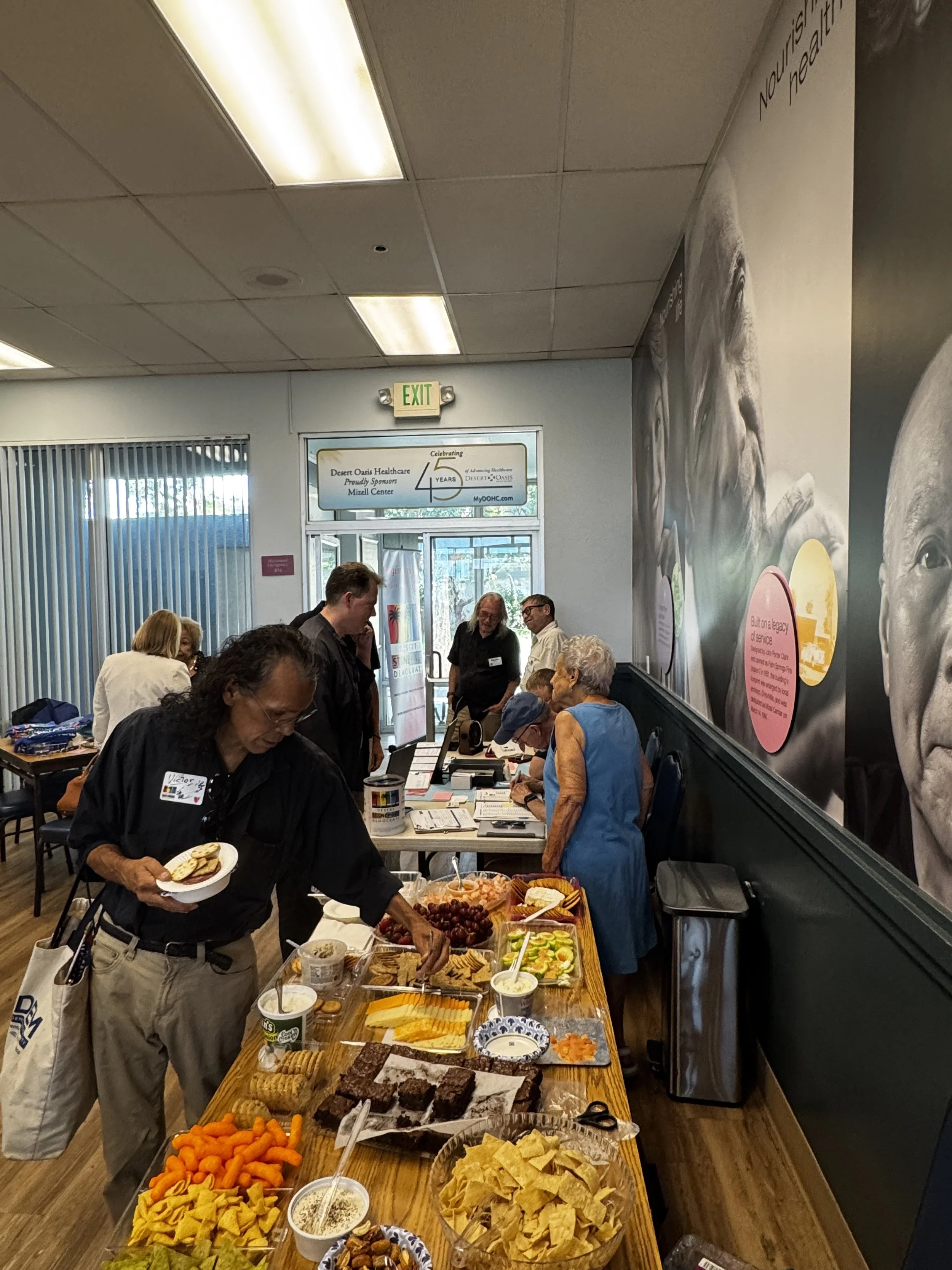 People gathered around a long table with various food items, including chips, vegetables, cheese, and desserts, at an indoor event or gathering.