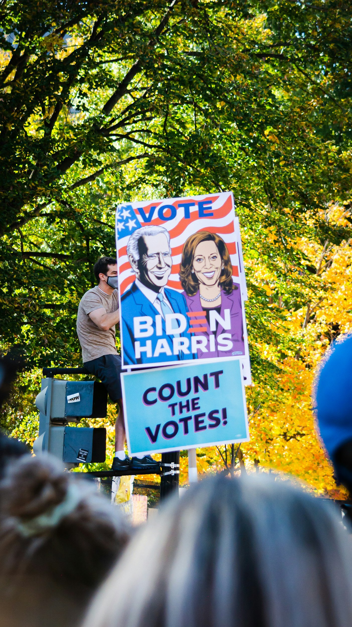 A person standing on a platform at a protest or rally holding a sign supporting Joe Biden and Kamala Harris with the words "VOTE BIDEN HARRIS" and "COUNT THE VOTES". The scene is outdoors with trees in fall colors in the background.
