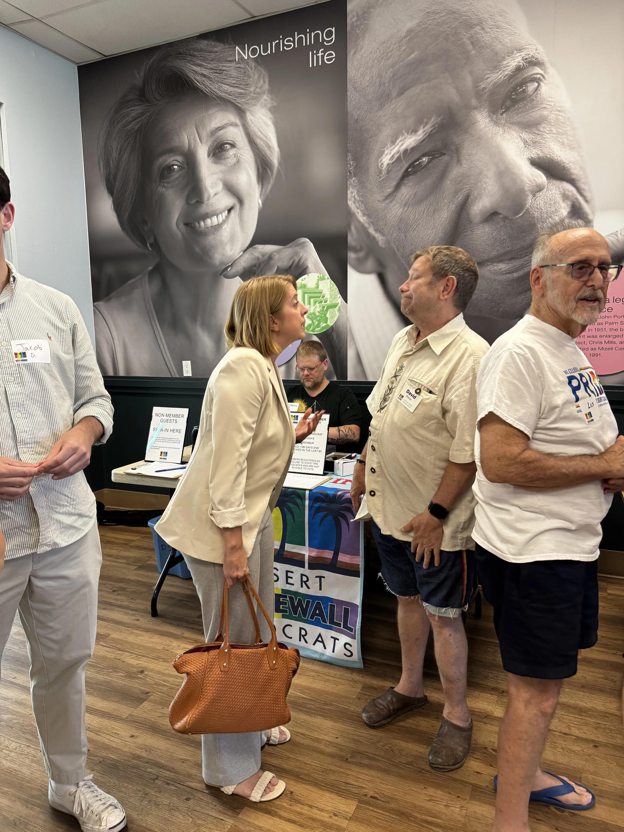 A group of five people standing and talking indoors, with large black and white portraits of two older individuals on the wall behind them. One woman with a tan blazer and light-colored pants is in conversation with a man with glasses, a white shirt,