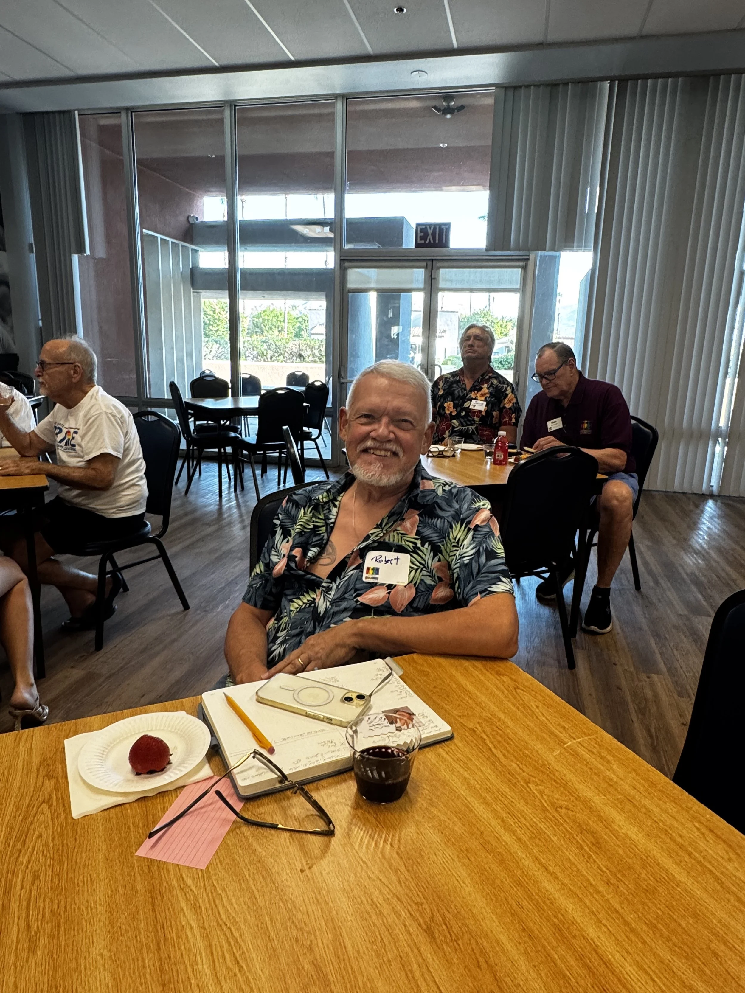 A group of people seated at a table in a room with large windows, enjoying a social gathering or celebration. The table has a plate with a red dessert, a glass of dark beverage, a notebook, and a pair of glasses. One man in the foreground is smiling 