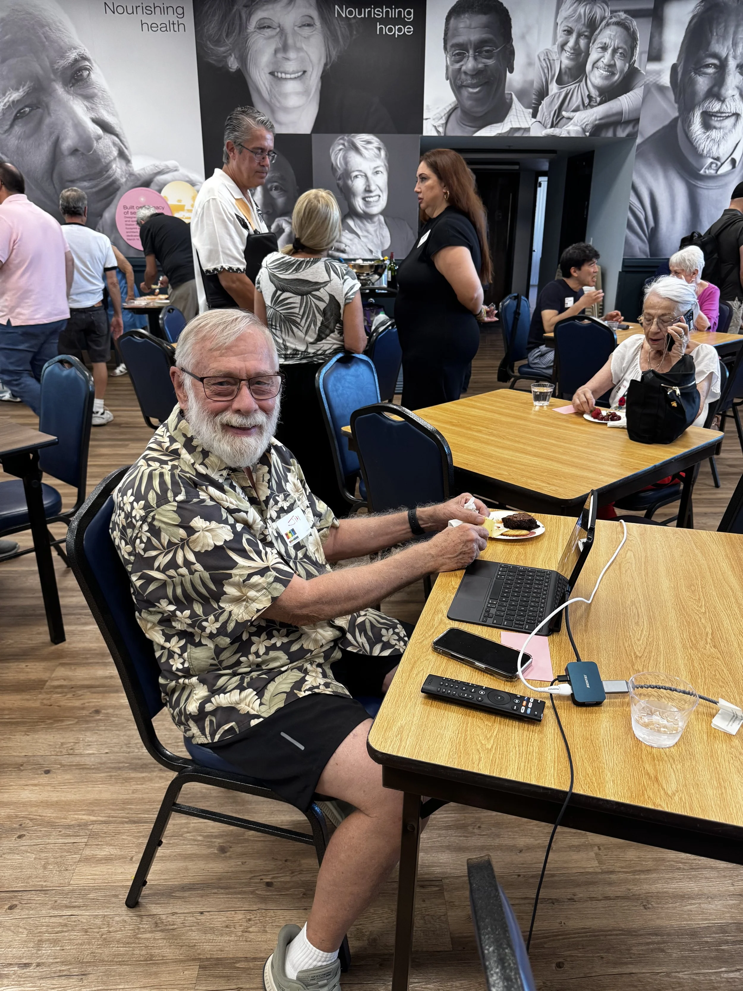 An elderly man with glasses, a white beard, and a floral Hawaiian shirt is sitting at a table in a conference or event space. He is smiling at the camera, with food on a plate in front of him and a laptop, remote, and phone on the table. In the backg
