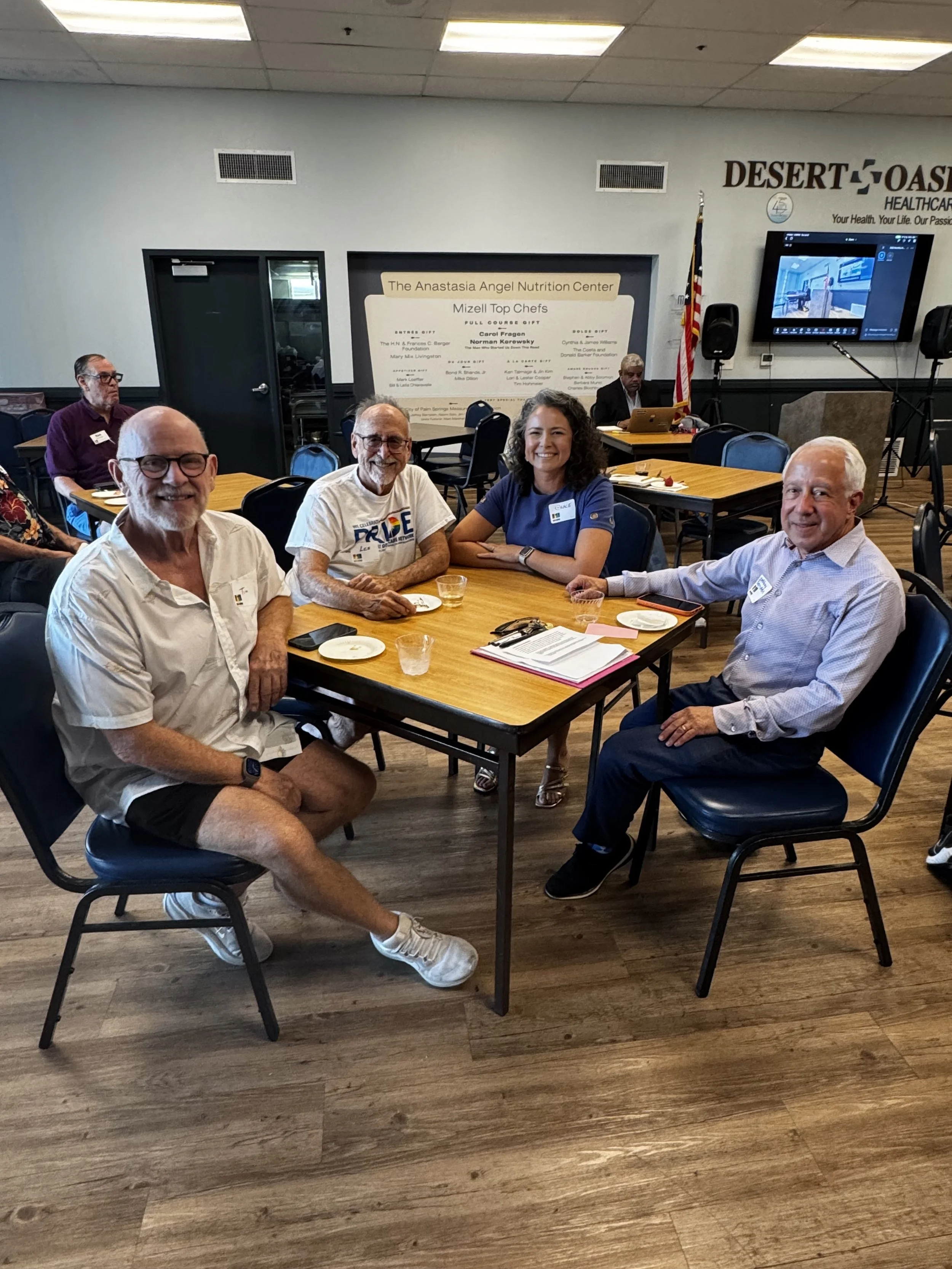 A group of five people sitting around a table in a conference room. The table has plates, cups, and documents on it. There is a large sign in the background that reads "The Anastasia Angel Nutrition Center Mizell Top Chefs," and a large digital scree