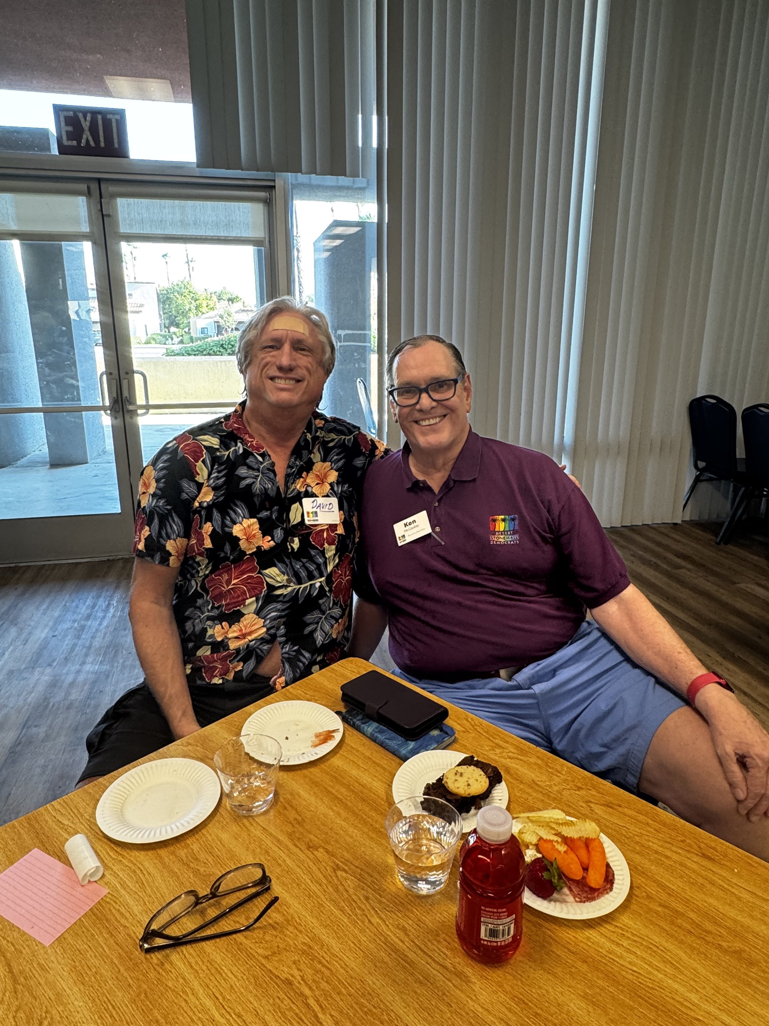 Two smiling men sitting at a table in a room with vertical blinds and glass doors, with partially eaten desserts, drinks, and personal items on the table.