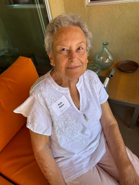 An woman with gray hair sitting on an orange cushion, wearing a white embroidered blouse and a name tag that reads Ruth. She is indoors, with a table behind her holding glassware and decor.