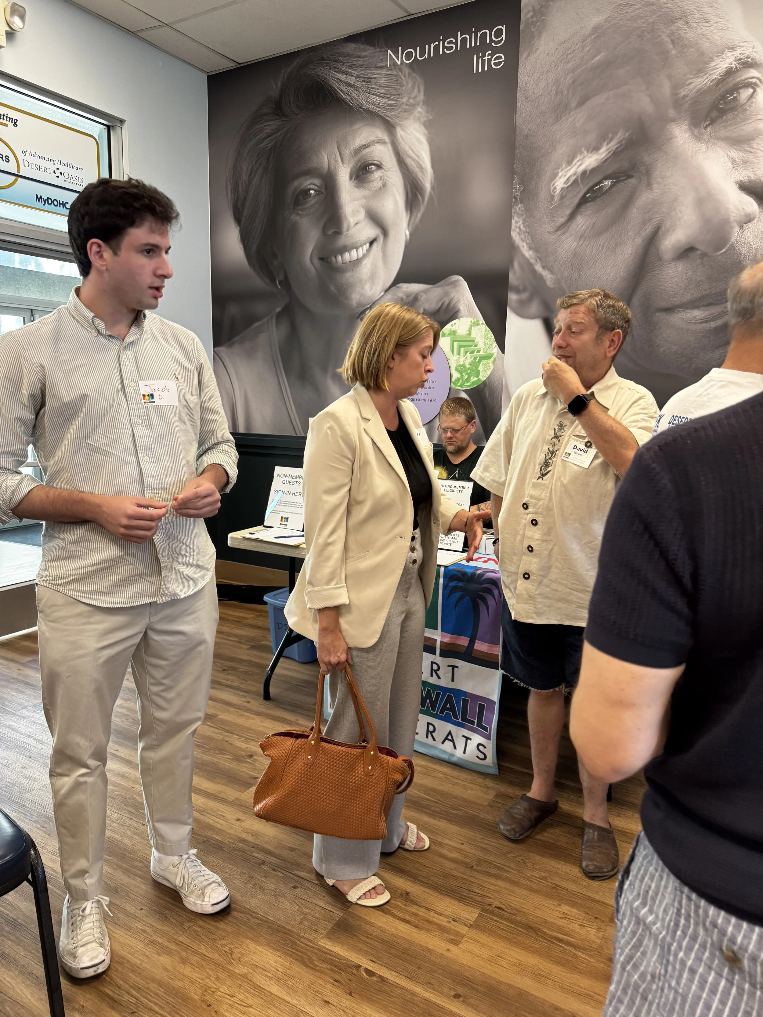 People gathered in an indoor setting, engaged in conversation, with a large black-and-white mural of a smiling woman and an elderly man on the wall behind them.