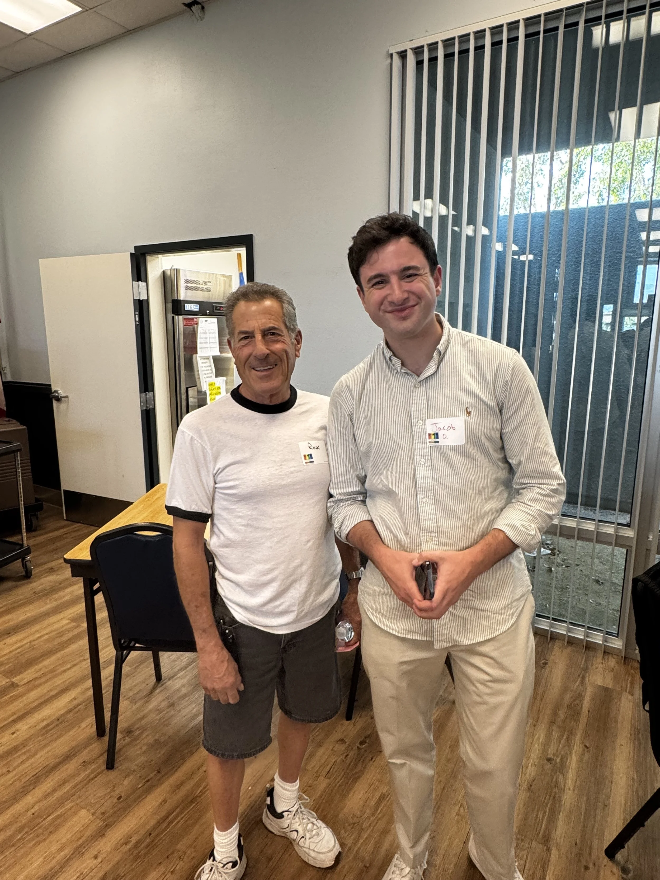 Two men standing side by side indoors, smiling at the camera. One is older with gray hair, wearing a white t-shirt with black trim and dark shorts, and the other is younger with dark hair, wearing a beige striped button-up shirt and light-colored pan