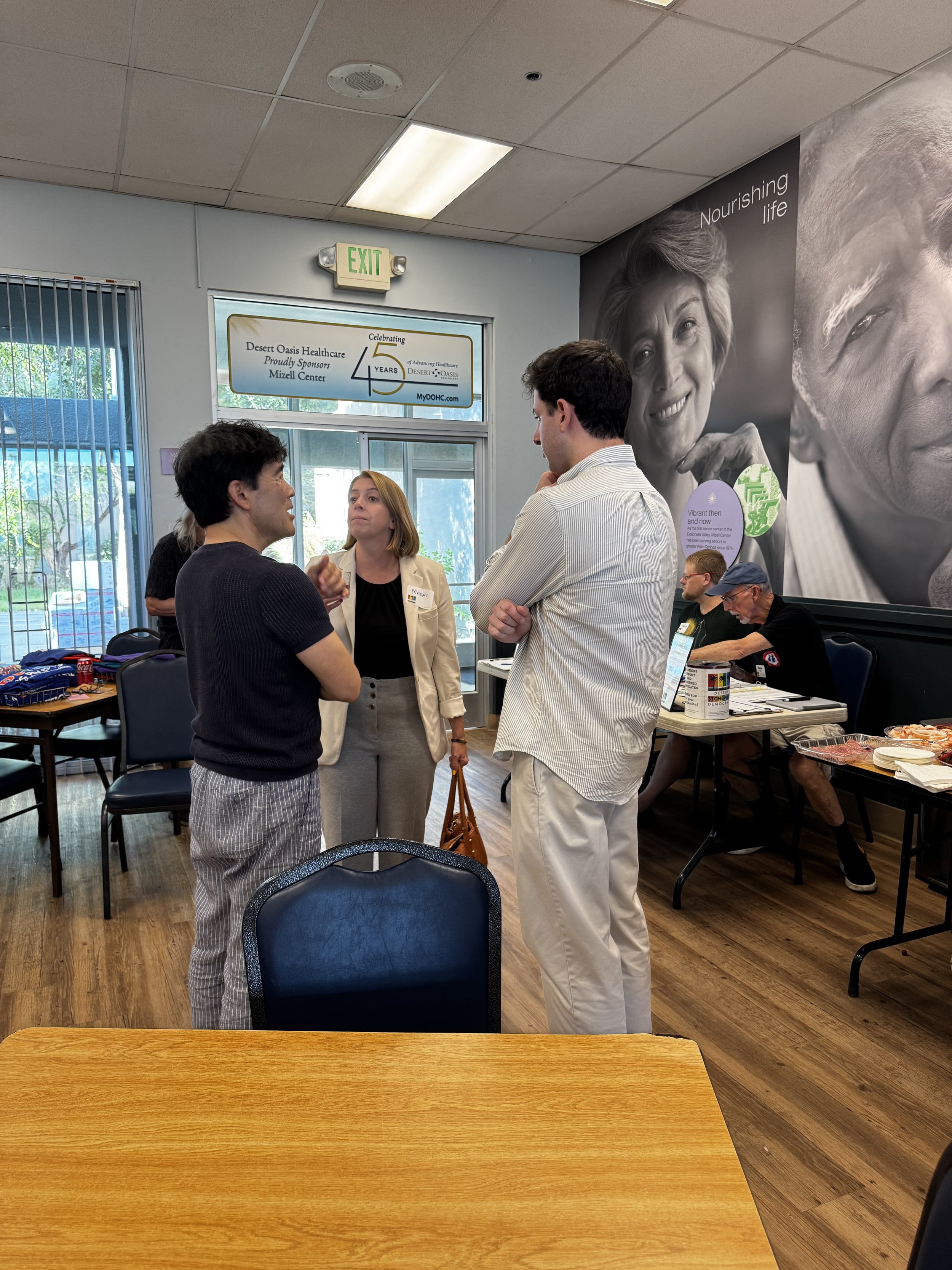 A group of four people engaged in conversation inside a room, with a large black and white mural of an elderly woman on the wall and a window with vertical blinds behind them.