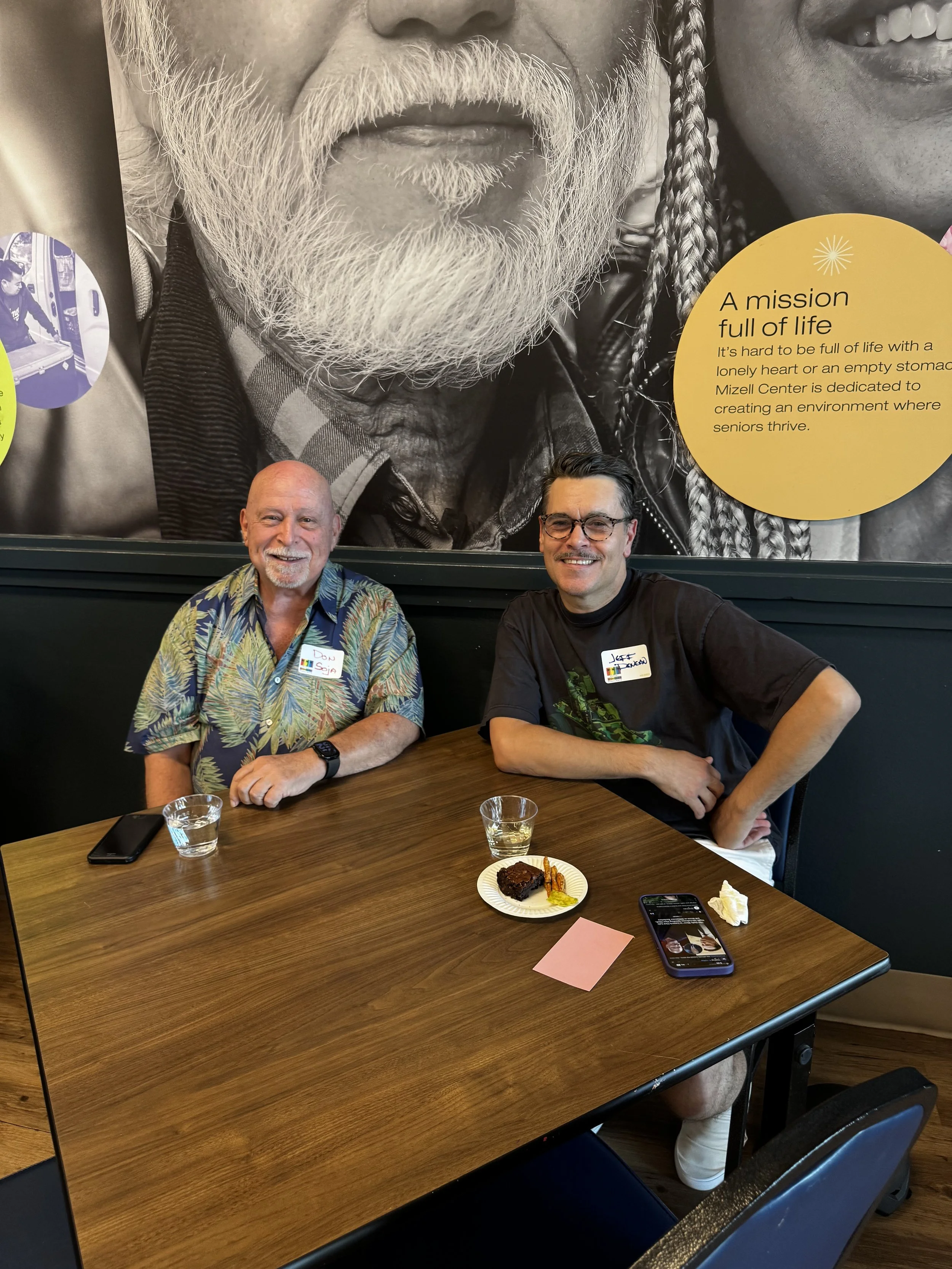 Two men sitting at a table with a small dessert and drinks, smiling and posing for the camera, with a large black-and-white mural of a bearded older man on the wall behind them.