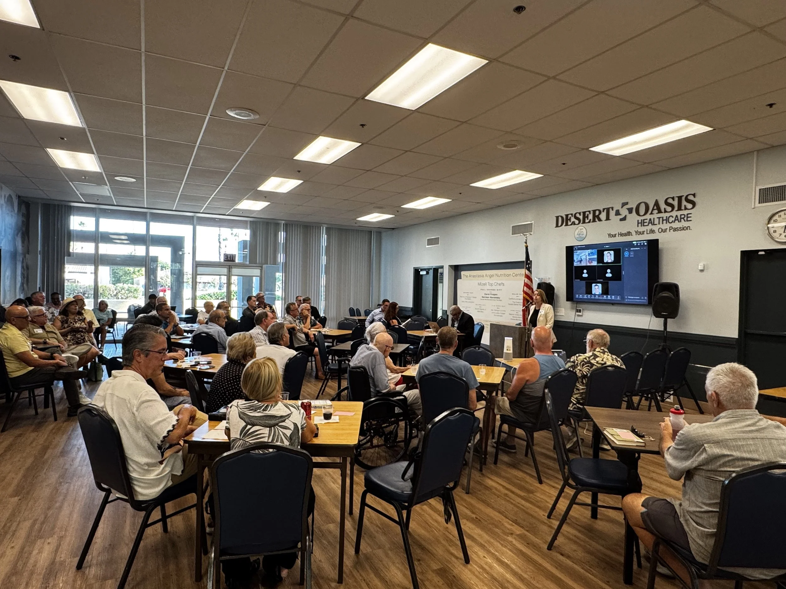 A large gathering of people seated at tables inside a conference room. There is a woman speaking at a podium in front, and a screen displaying a video call. The room has large windows, a logo on the wall reading "Desert Oasis Healthcare," and a clock