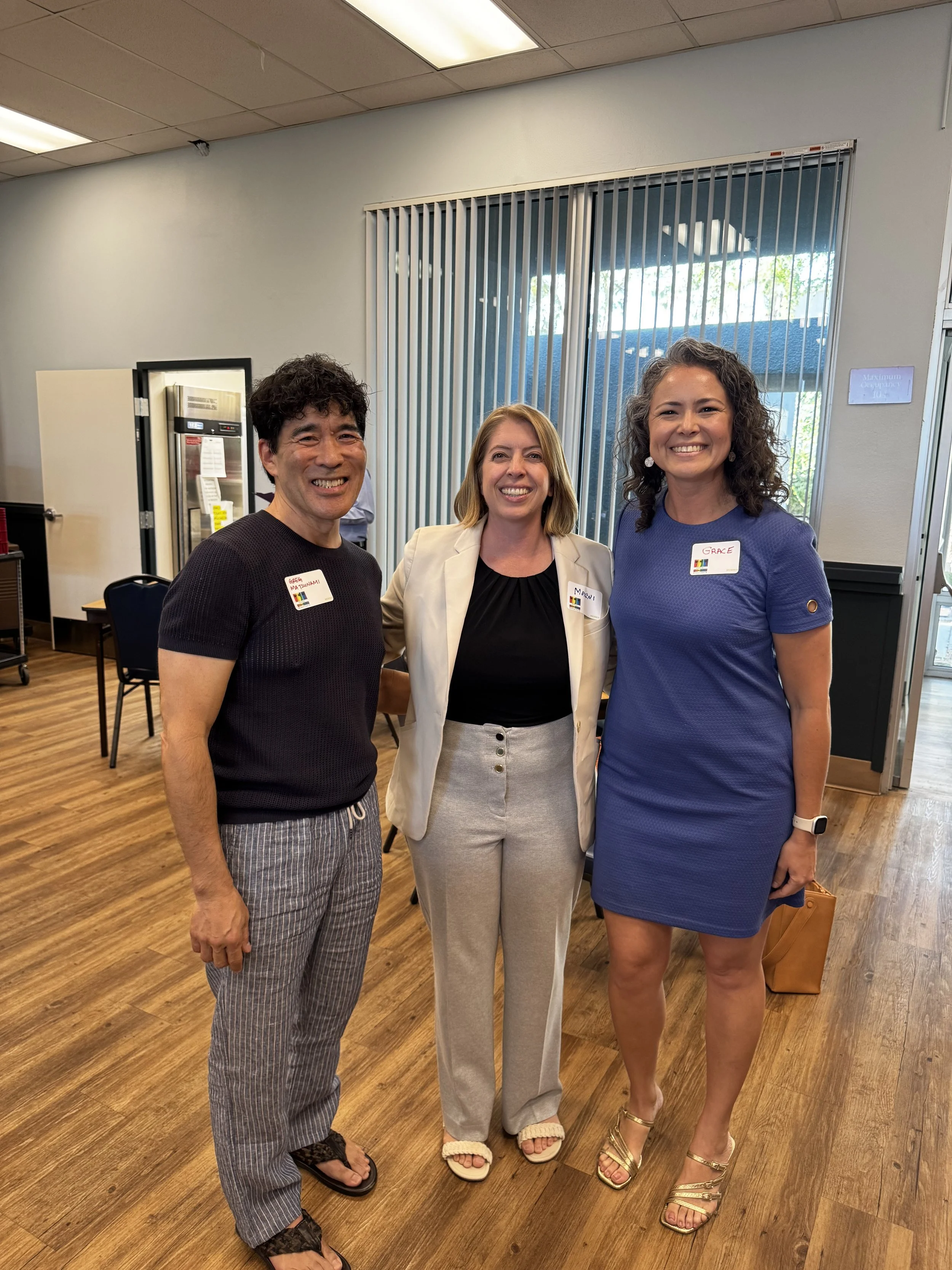 Three women standing together in an indoor setting, all smiling and wearing name tags, with a window and vertical blinds in the background.