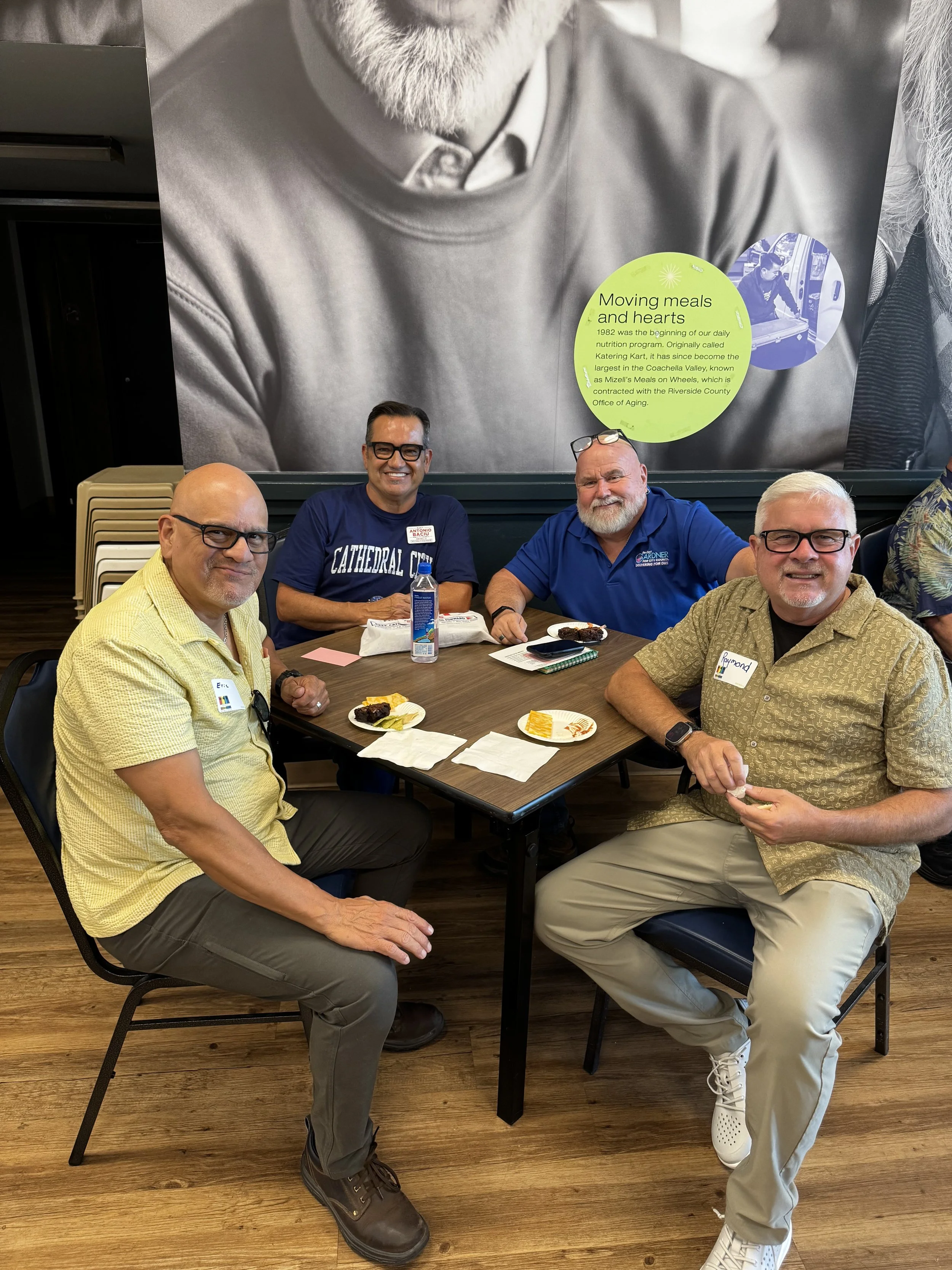 Four men sitting around a table enjoying a meal, with plates of food and drinks in front of them. They are smiling and appear to be in a friendly gathering.