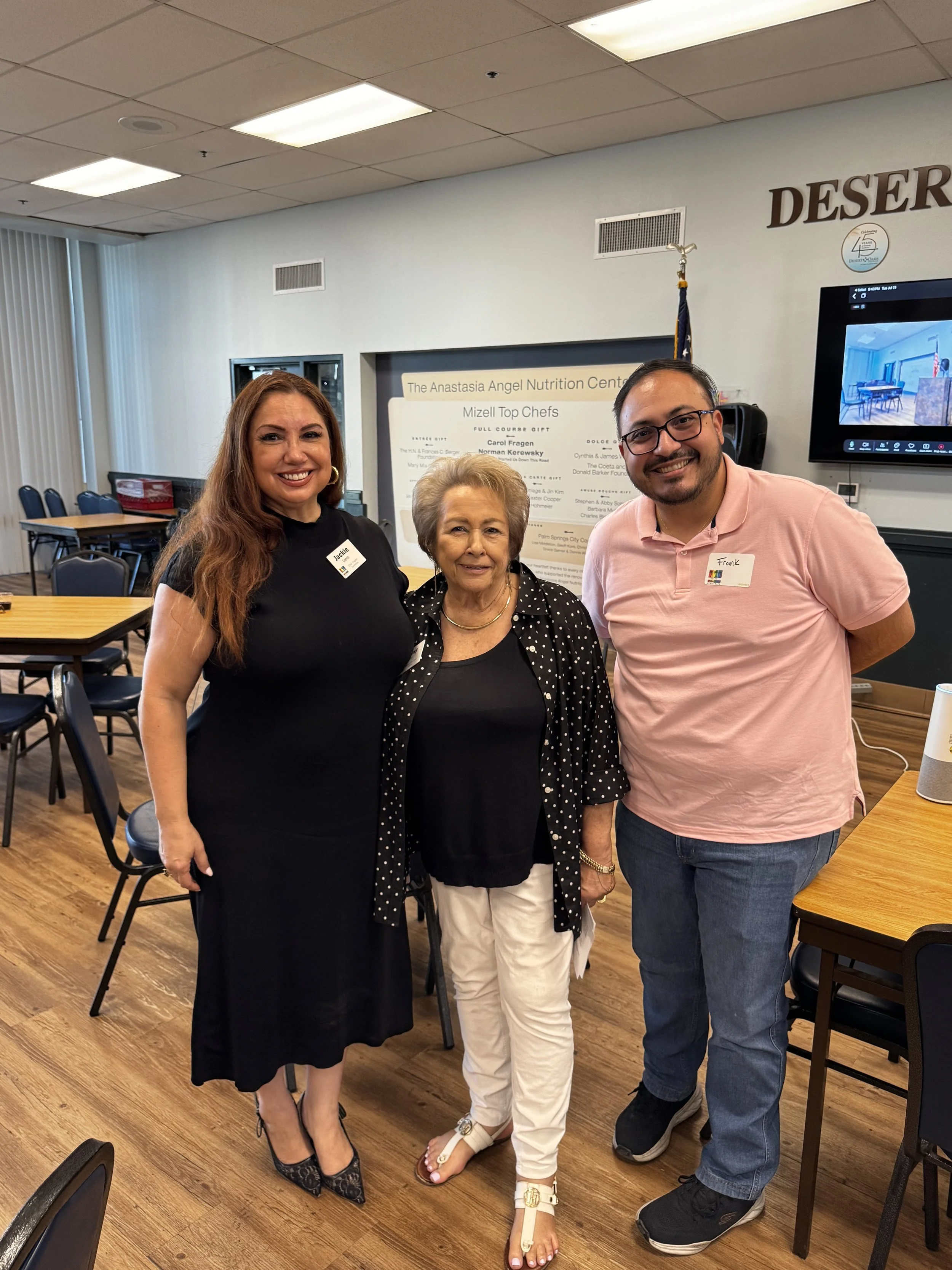 Three women standing together inside a conference or event room with tables and chairs, smiling at the camera. They are dressed in semi-formal attire, with name tags on their shirts. A sign on the wall behind them mentions the Anastasia Angel Nutriti