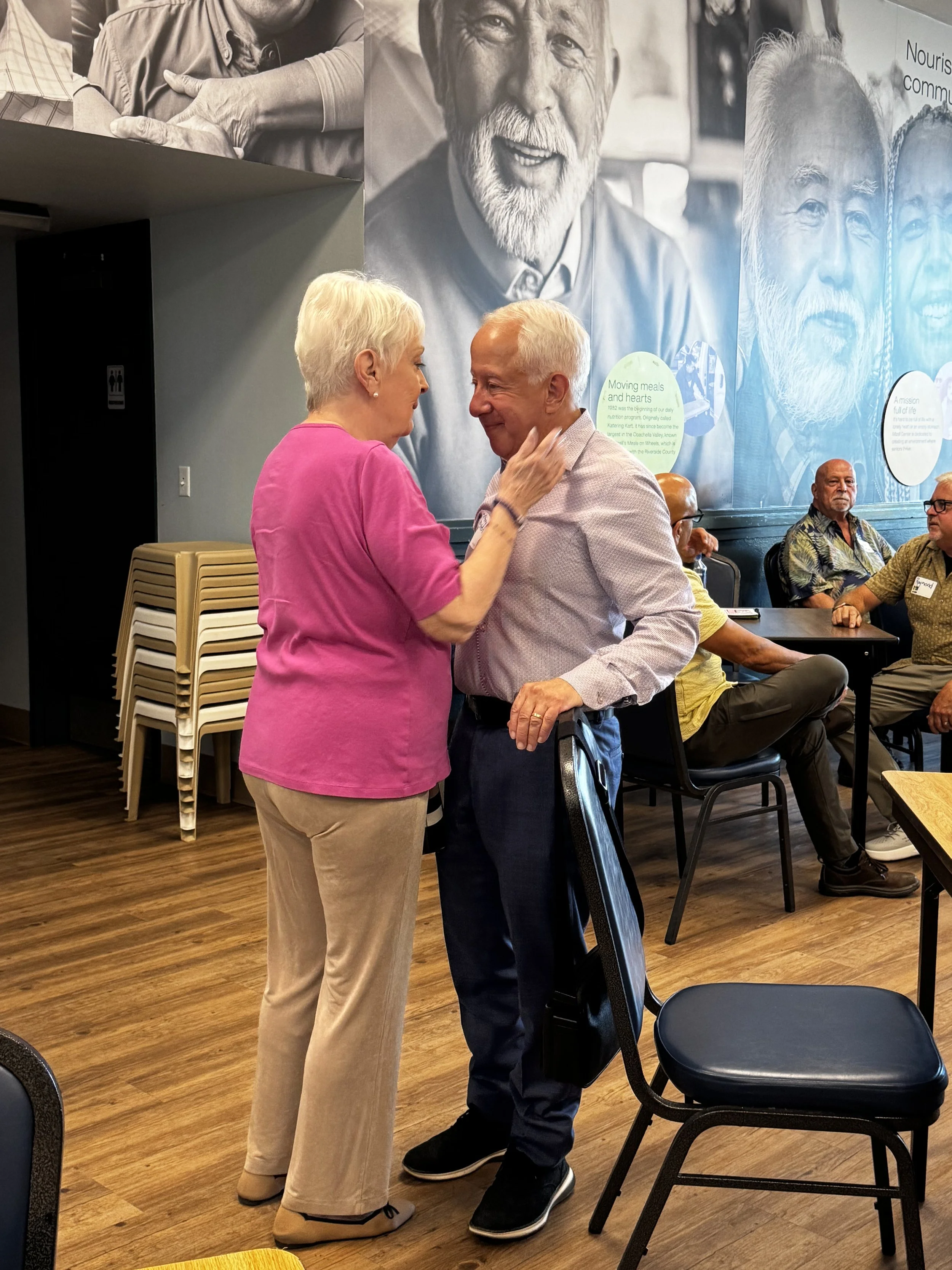 An elderly woman in a pink top and beige pants touching foreheads with an elderly man in a light shirt and dark pants in a social setting. Several people are sitting at tables in the background, and large black-and-white portraits are displayed on th
