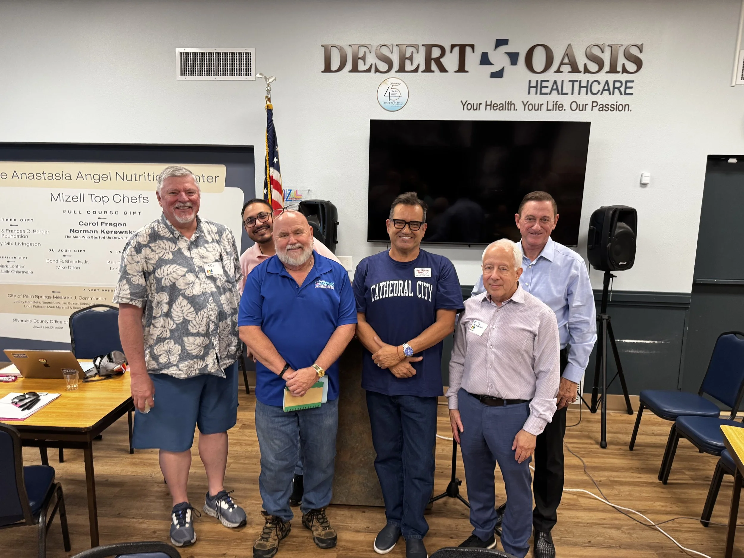 A group of seven men standing together in a room with a sign that says 'Desert Oasis Healthcare' on the wall behind them. They are smiling and posing for the photo. The room has chairs, tables, and a large monitor on the wall.