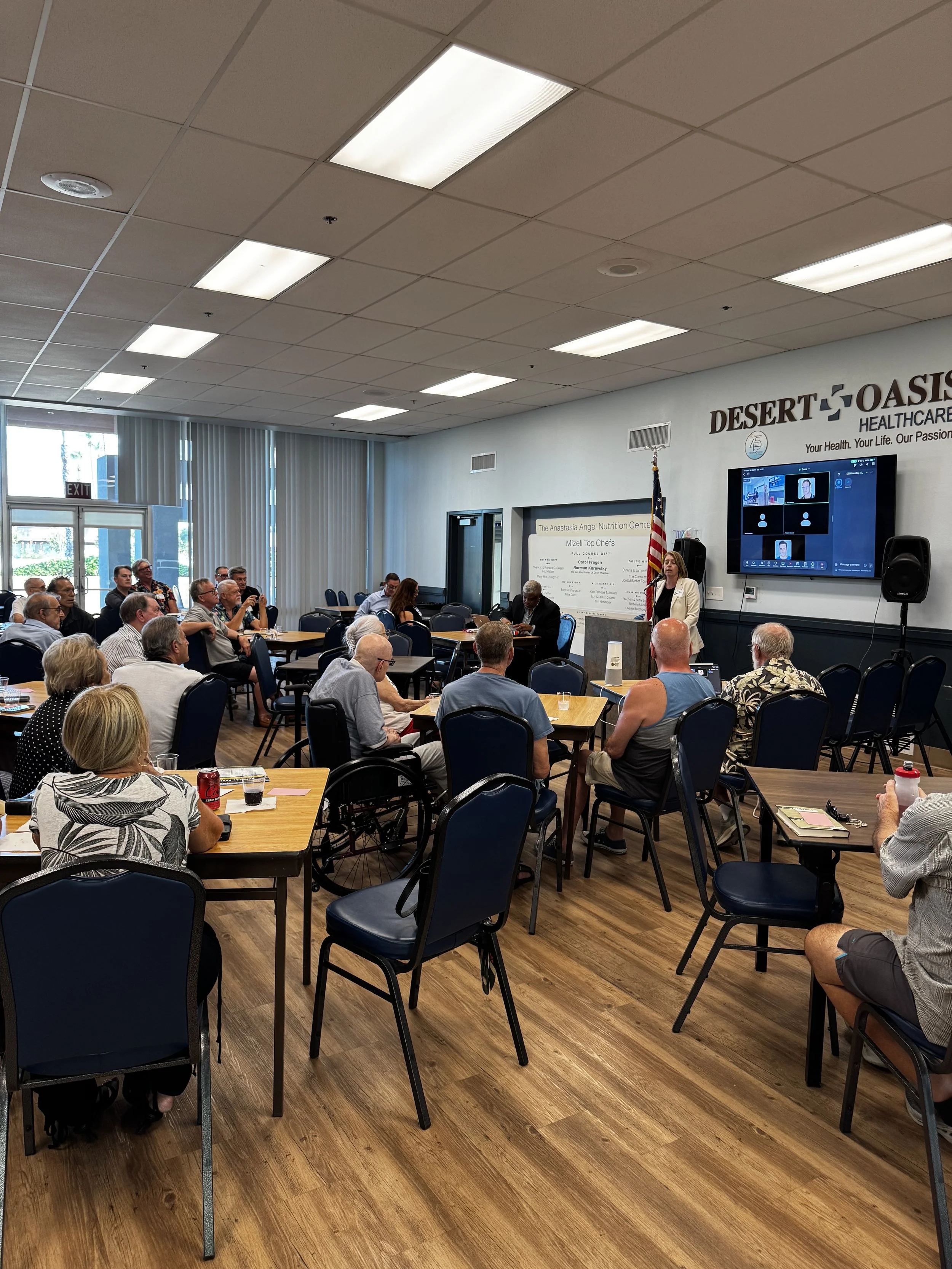 A group of people attending a presentation or meeting in a conference room with wood flooring, tables, chairs, a large screen on the wall, and an American flag. A woman is speaking at a podium near the screen.