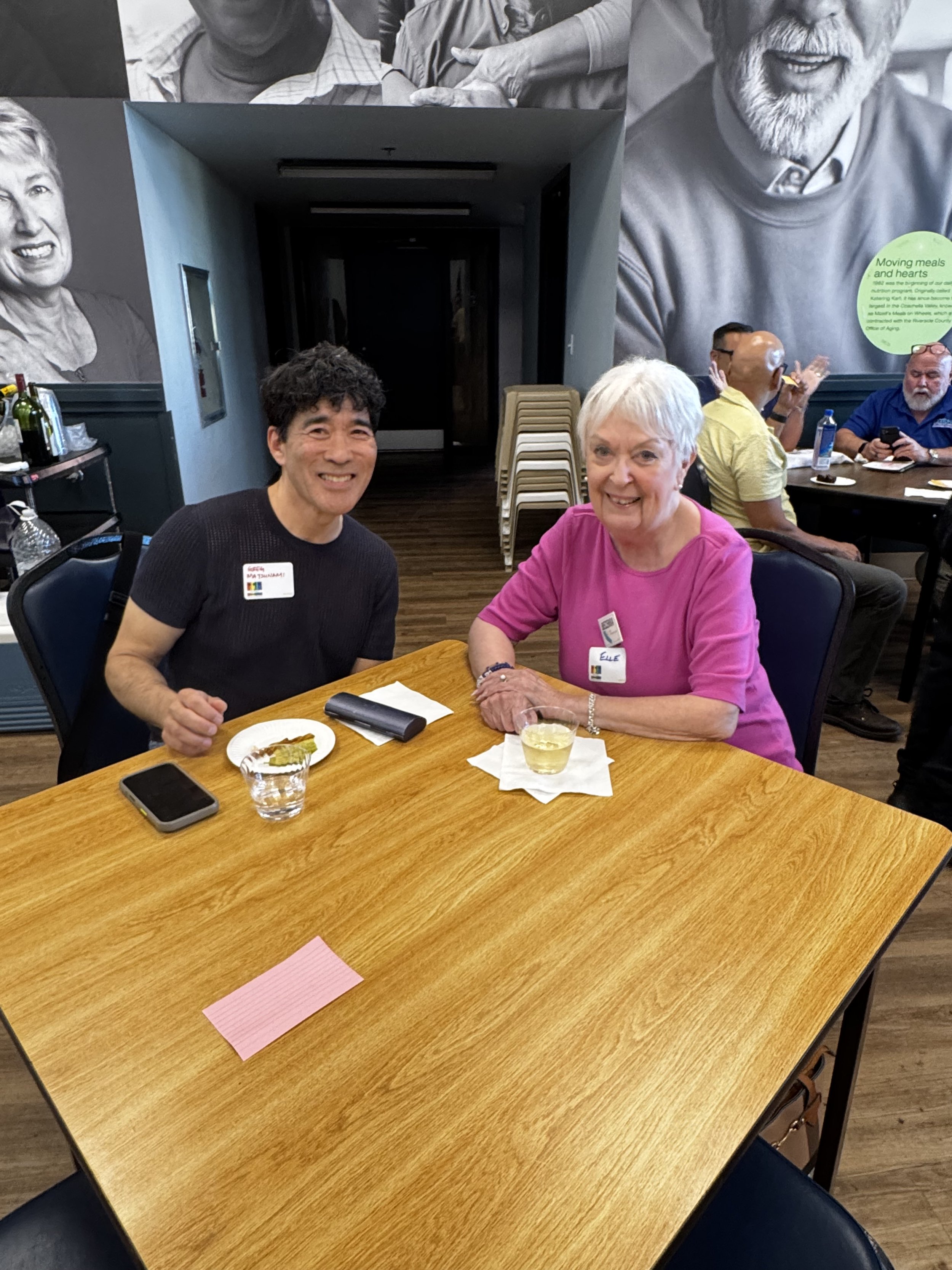 Two smiling women sitting at a wooden table with drinks and a small piece of food, in a room with people in the background, some of whom are clapping, and large black-and-white photographs of older adults on the wall.