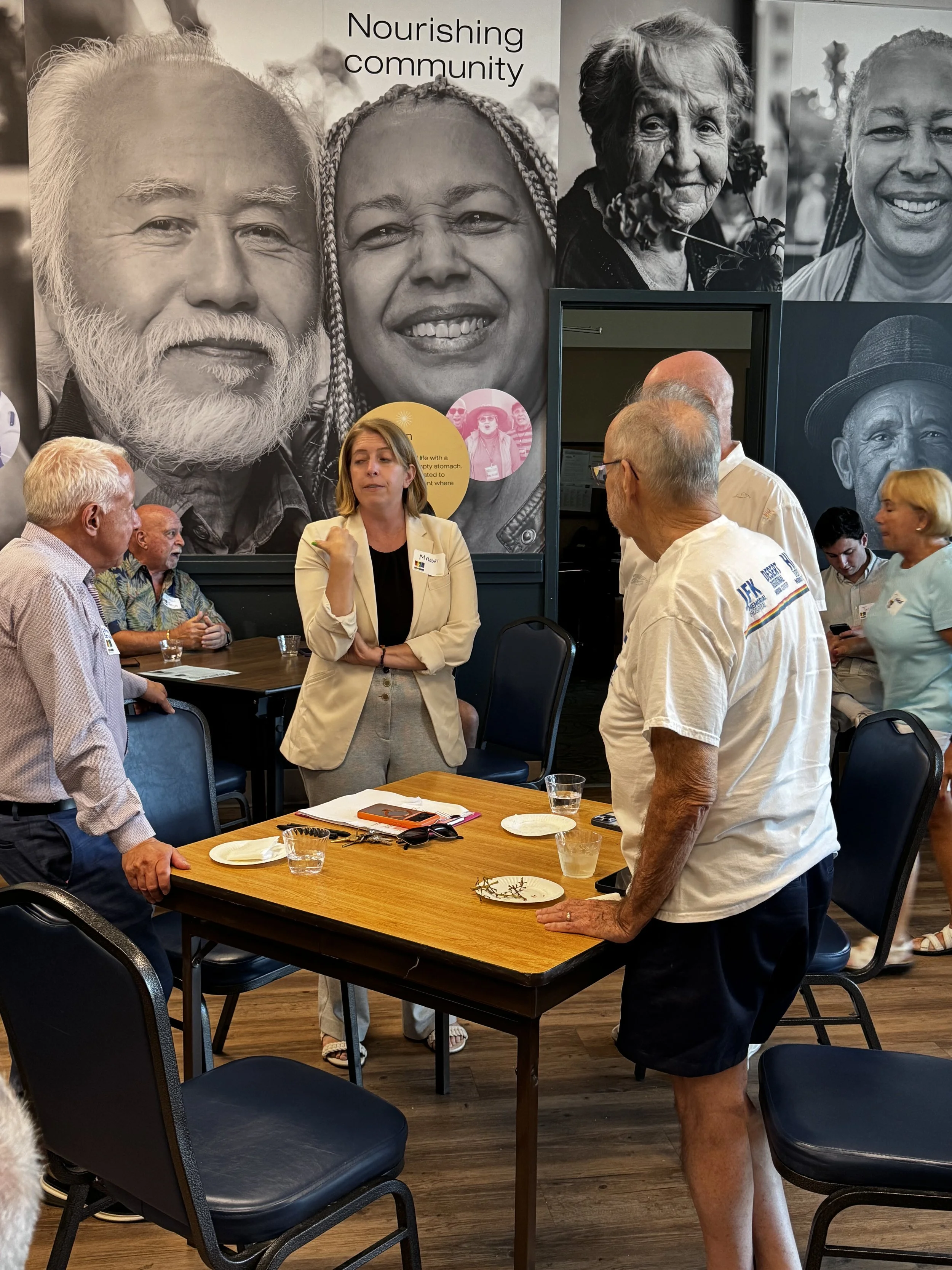 A group of people having a discussion in a room decorated with a large black-and-white mural of diverse smiling faces, including an elderly Asian man with a white beard, a smiling Black woman with braided hair, and other smiling individuals.
