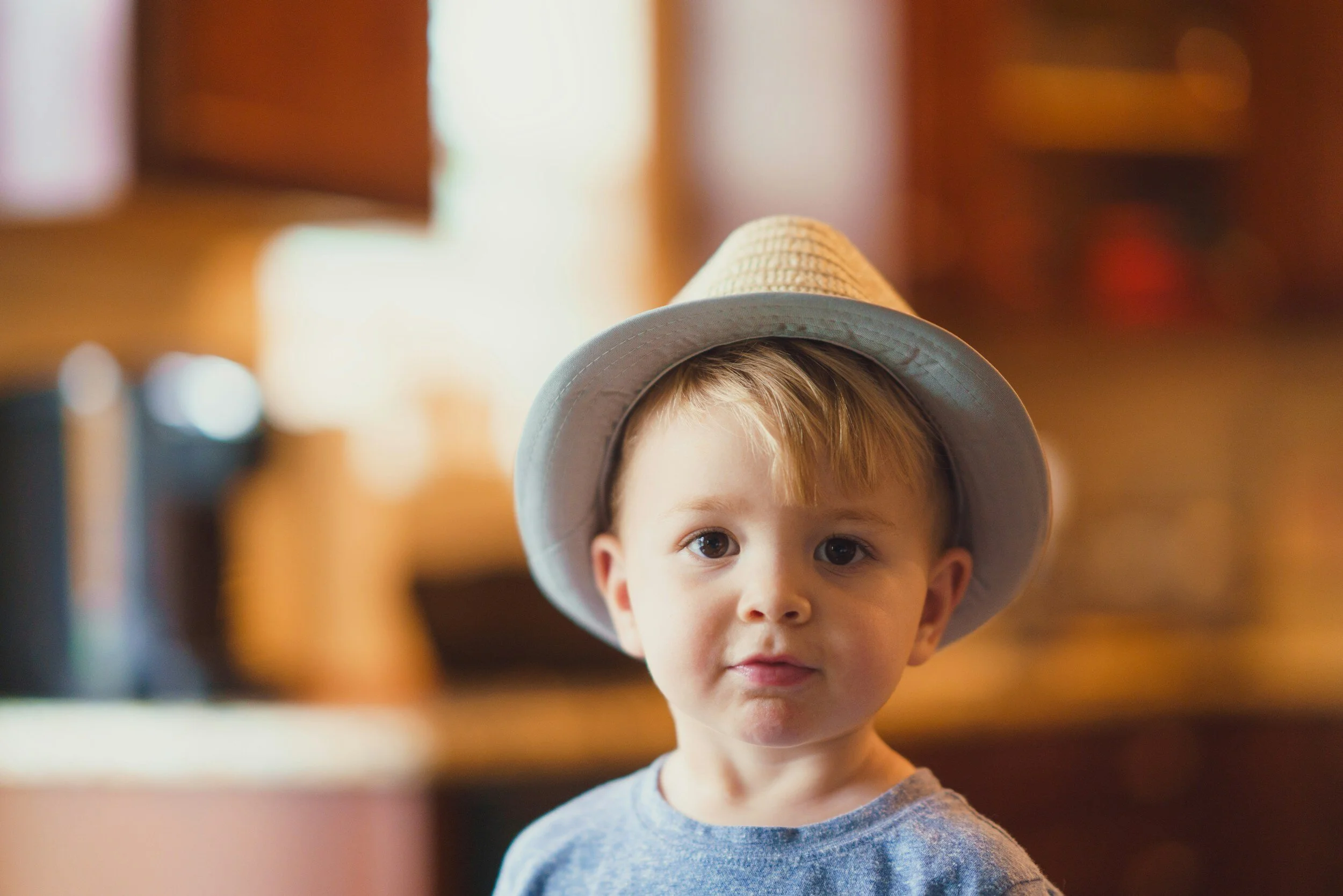 Young boy wearing a light-colored fedora hat and a gray shirt, indoors with warm lighting and blurry background.