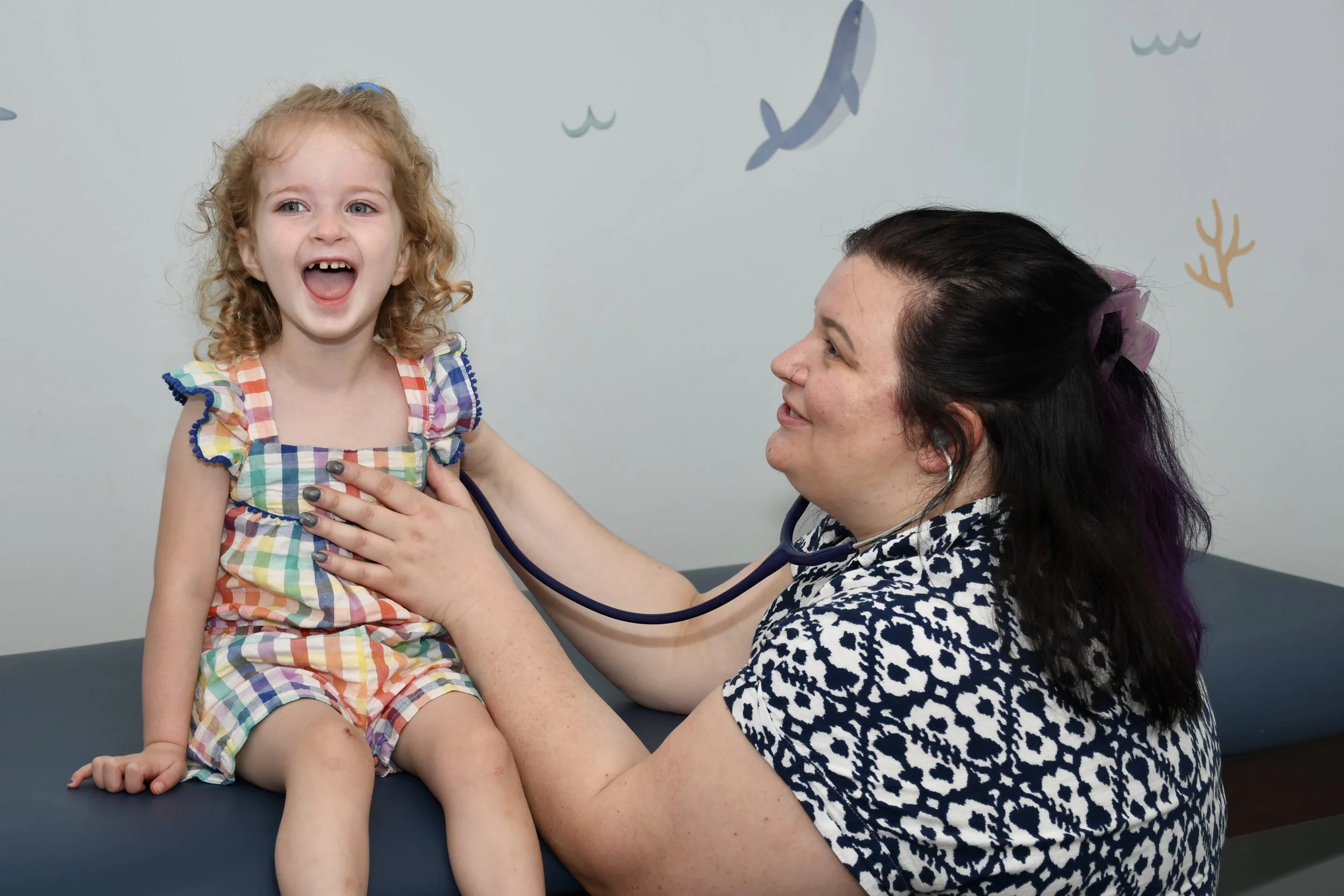 A young girl and female doctor in a pediatric clinic, with colorful handprints and the phrase "The Future of the World is in This Room" on the wall in the background.