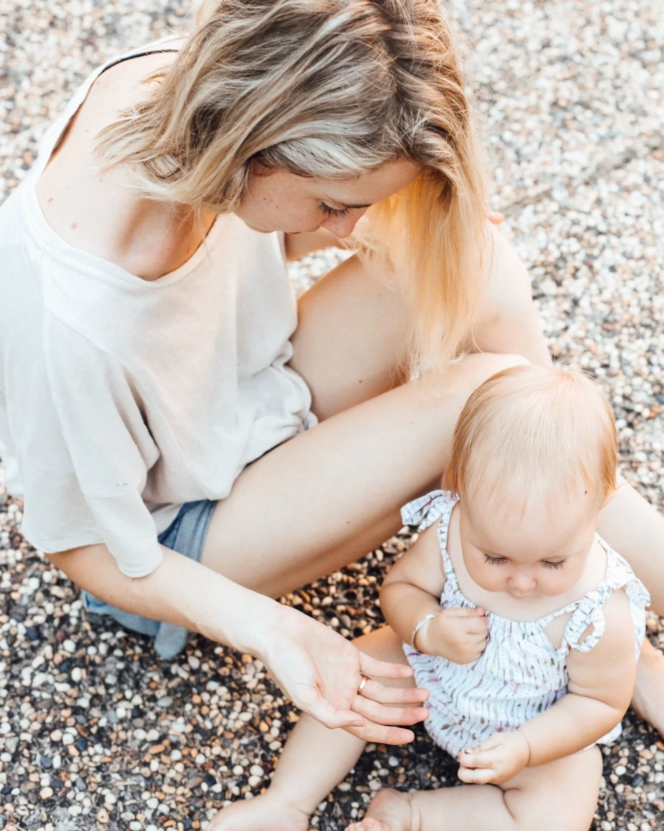 A young woman sitting on a pebble beach with a toddler, looking down, with the toddler sitting on her lap and looking down, both with light hair, the woman wearing a white shirt and shorts, and the toddler wearing a striped dress.