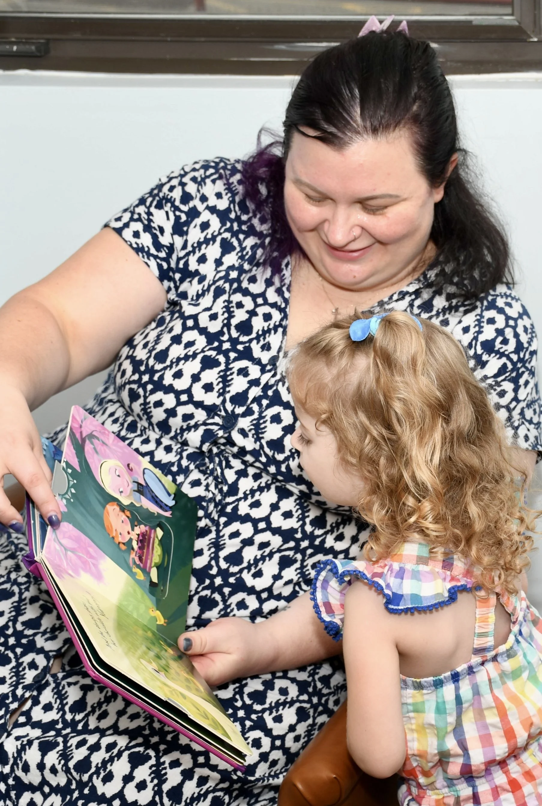 A woman and two young girls sitting on a colorful rug inside, engaging with a children's book and toys, while another woman sits in the background smiling.