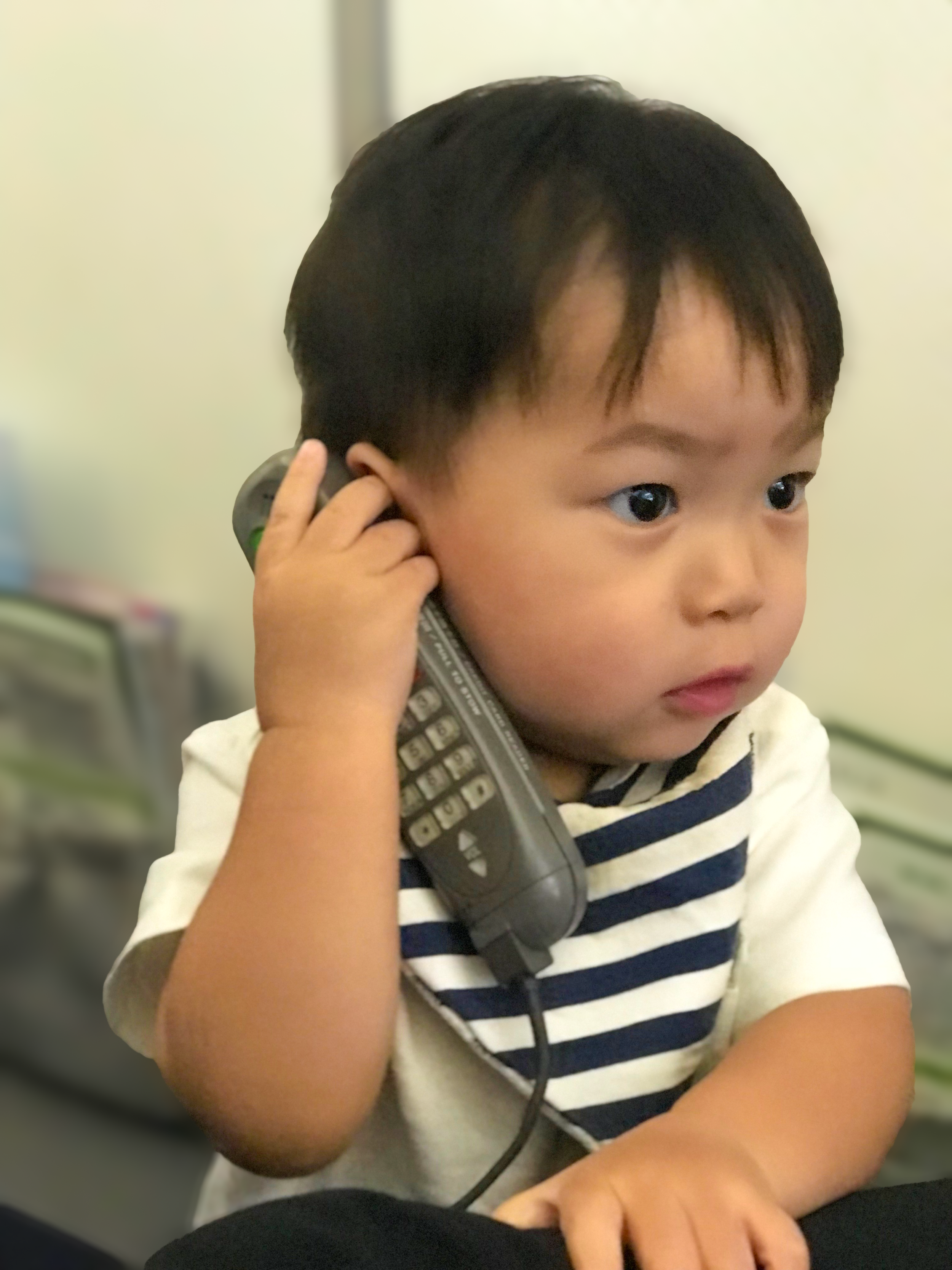 A young child with a rainbow headband and colorful sweater, holding a toy telephone, smiling at the camera inside a home.