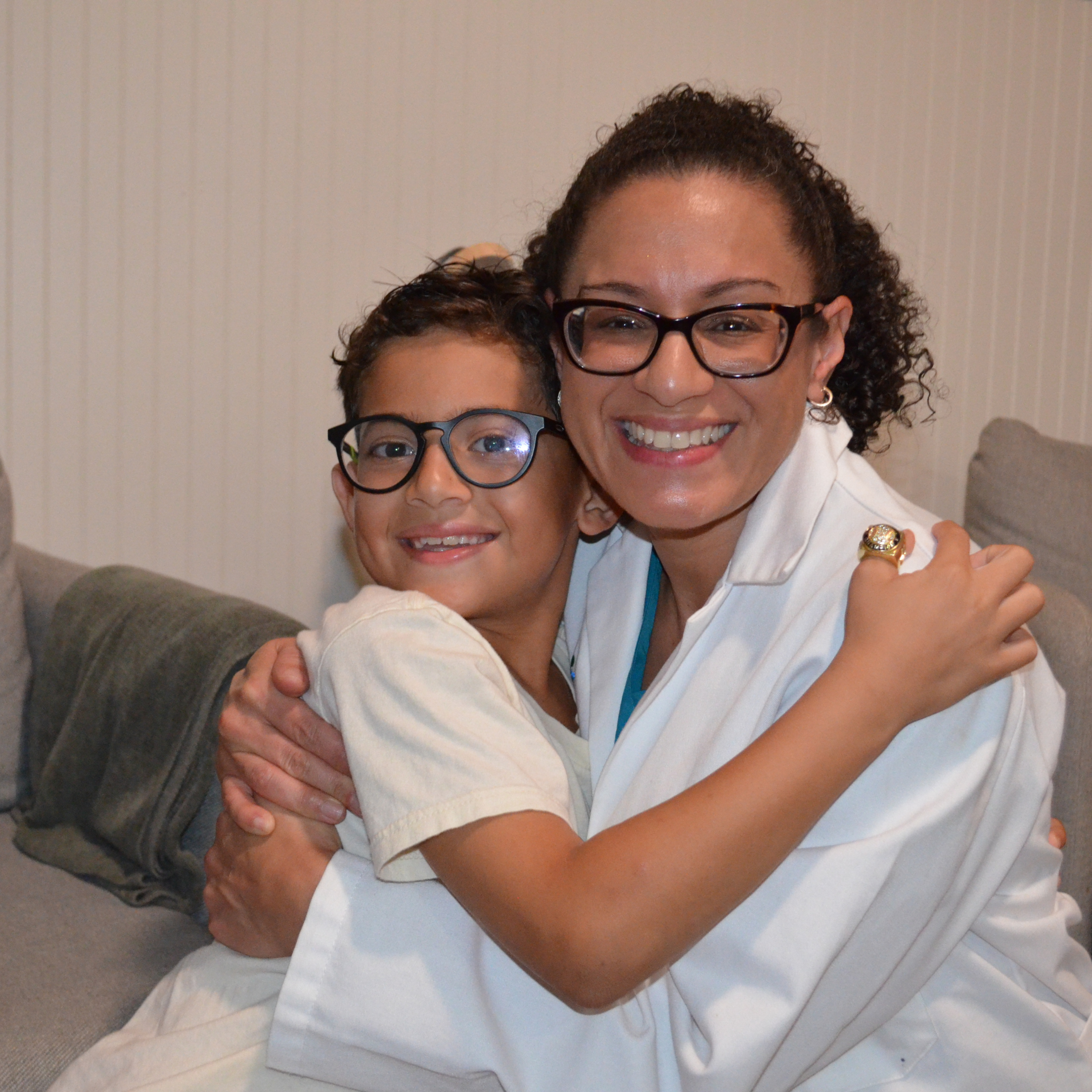 A healthcare worker kneeling next to a young girl in a colorful dress, both holding vaccination records and smiling; the worker is wearing medical scrubs and a face mask, indoors in a clinic or office.