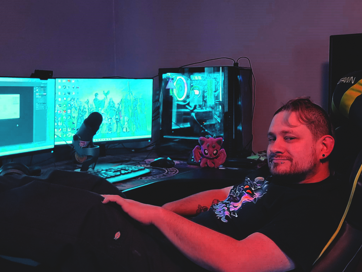 A man sitting in a gaming chair in front of dual computer monitors, with a gaming PC and plush toy on the desk, in a dimly lit room.