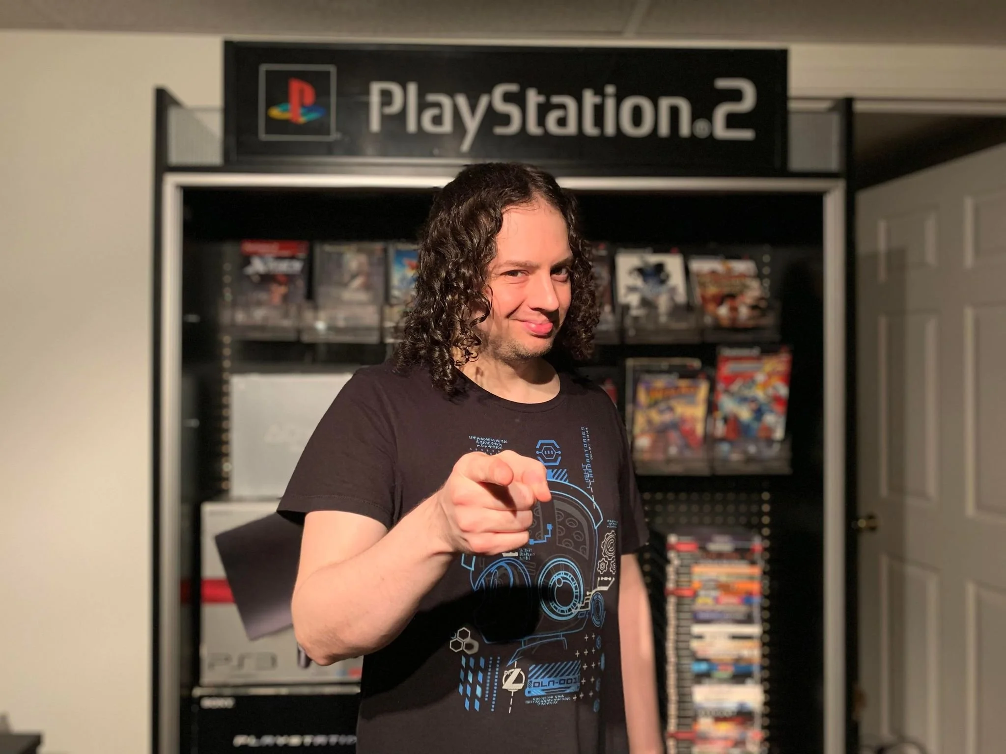 Person with long, curly hair pointing at the camera, standing in front of a display of PlayStation 2 games.