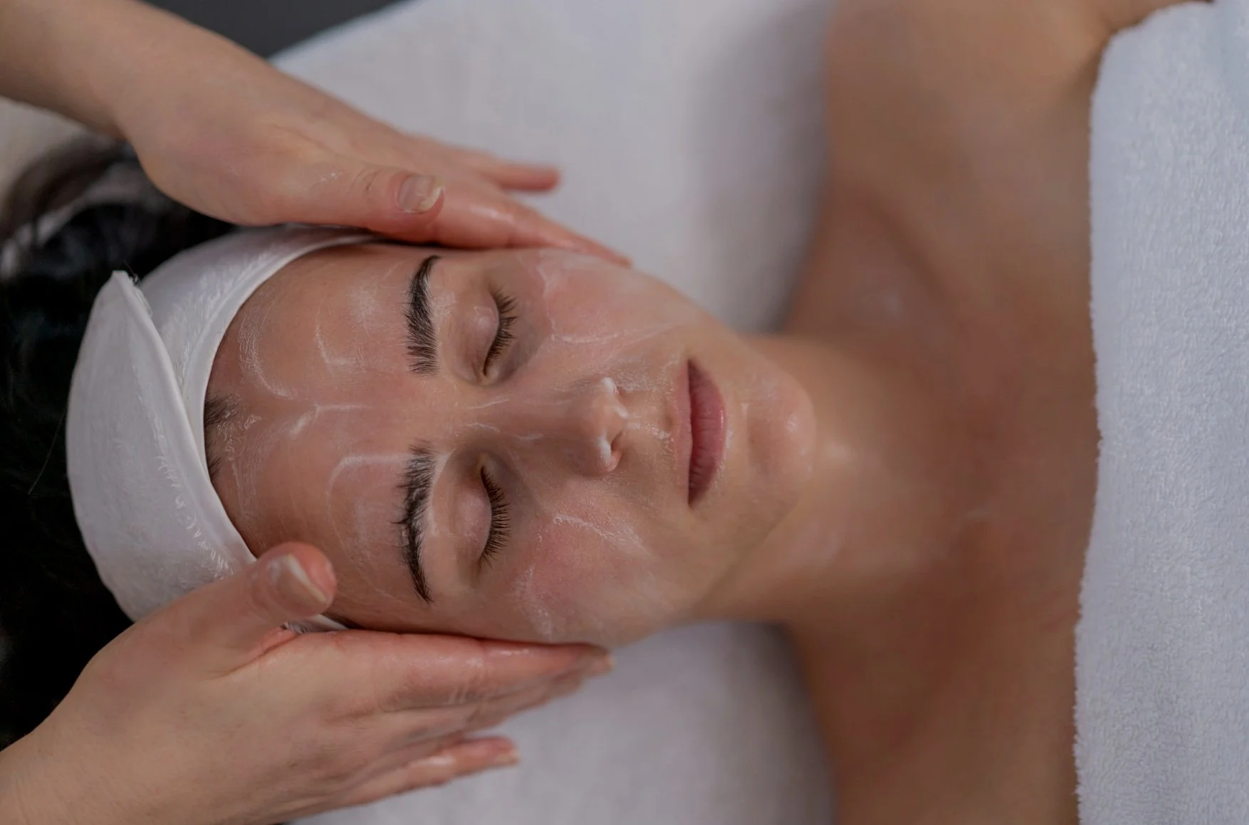 Woman receiving facial massage at spa, lying with eyes closed, head wrapped in a white towel.