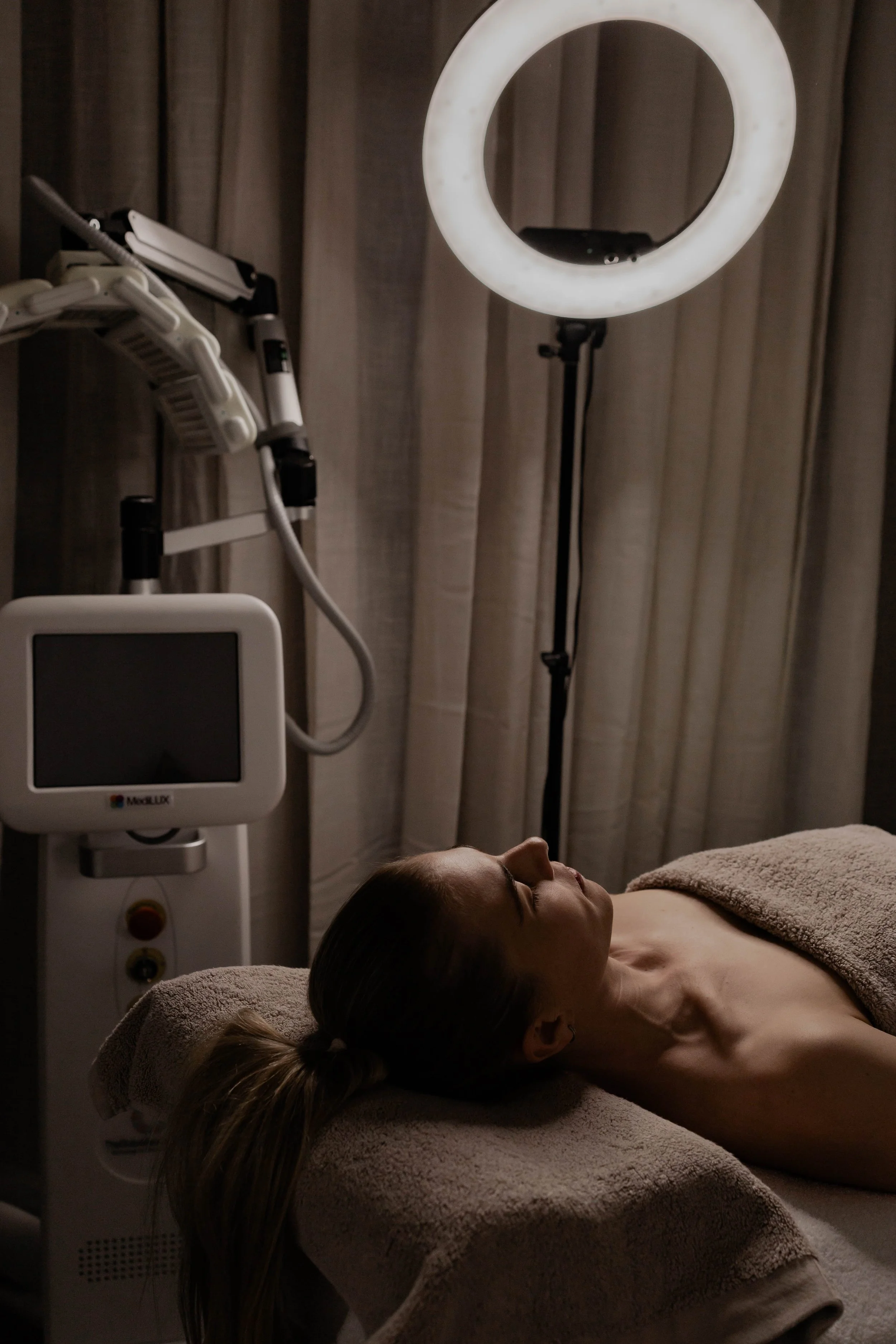 A woman lying on a massage table in a dimly lit spa or medical treatment room, with her eyes closed and a towel covering her lower body. There is a ring light and LED machine nearby, and curtains in the background.